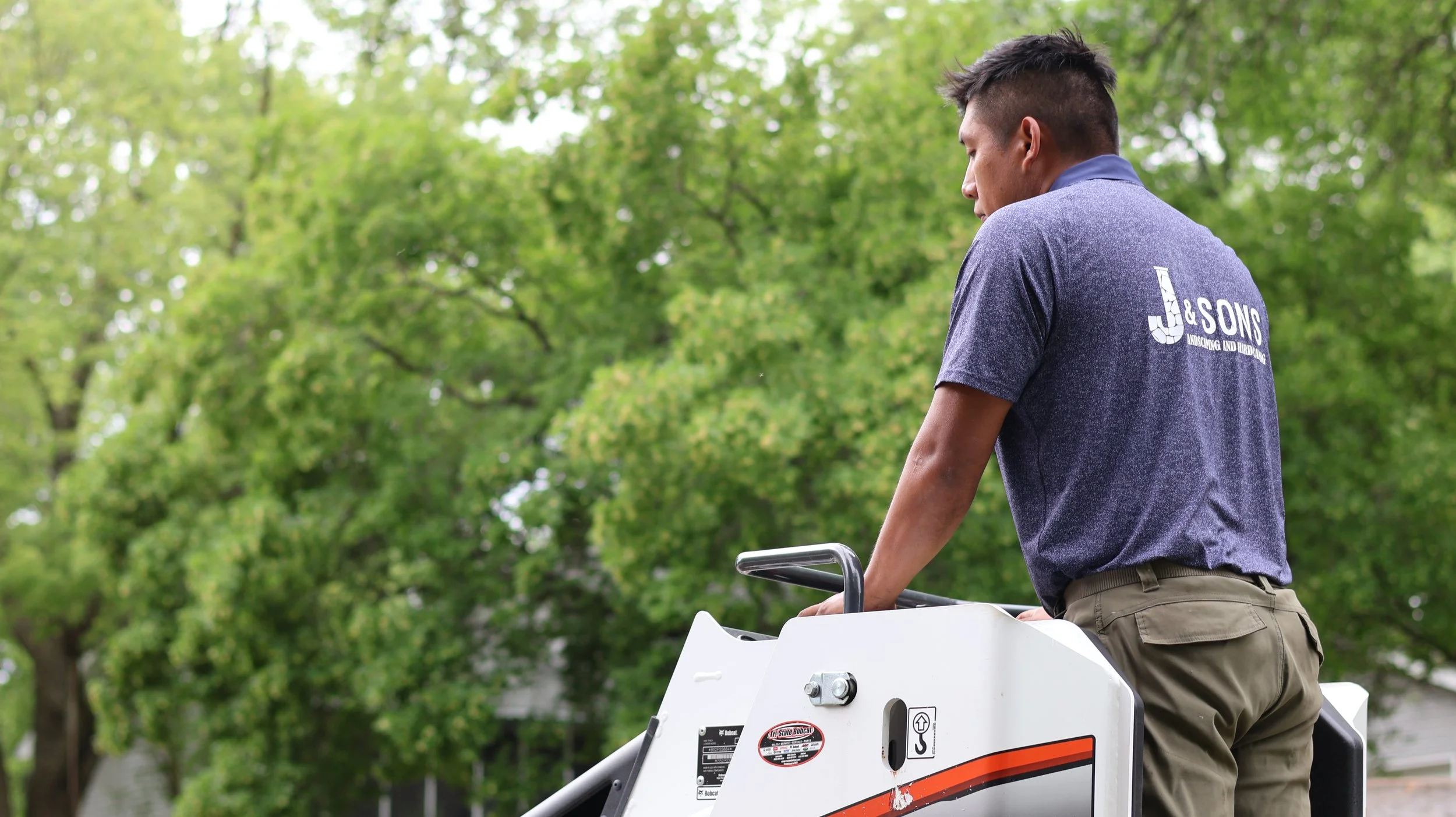A man riding a white Bobcat utility vehicle outdoors among green trees.