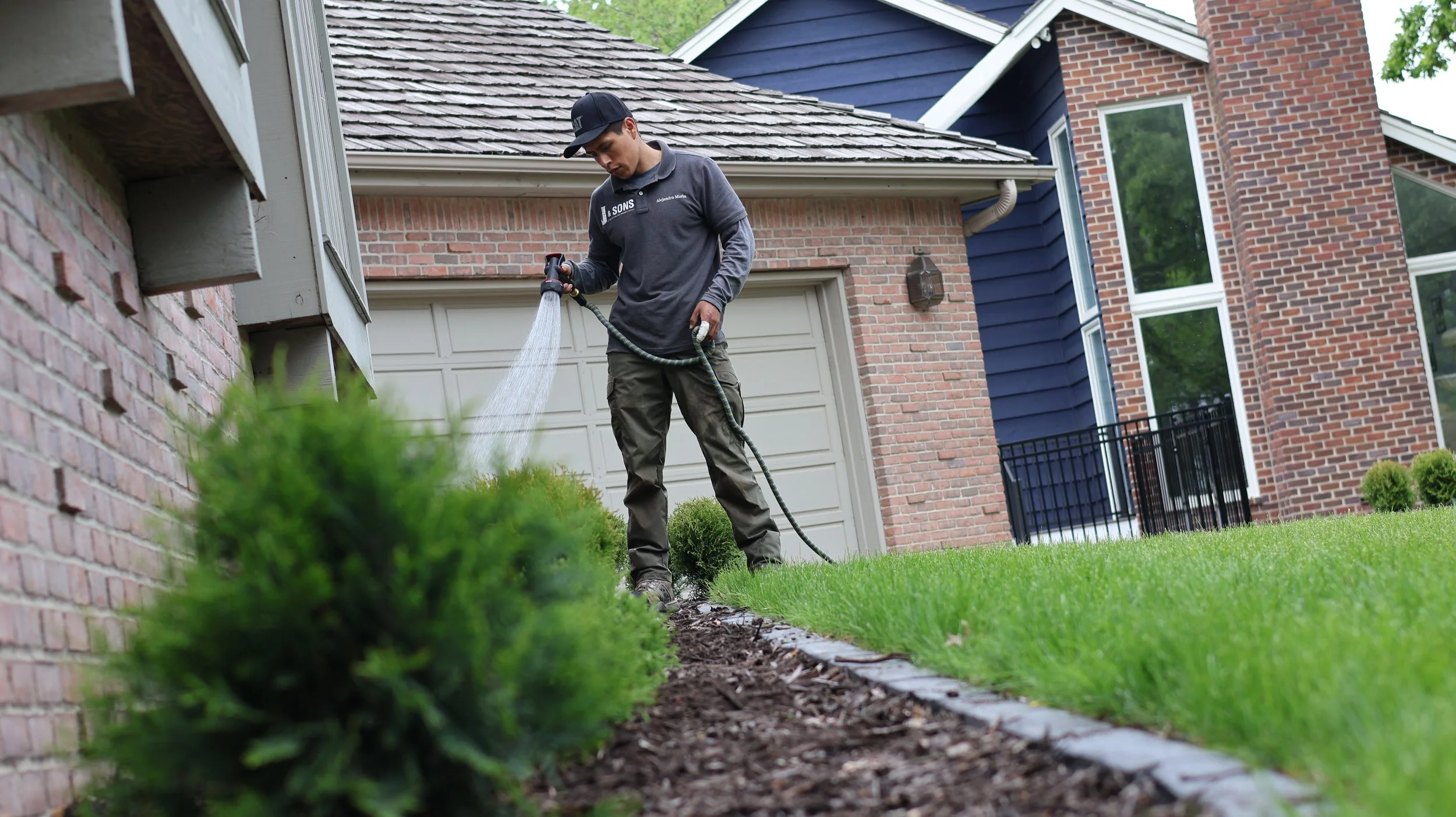 A person watering a garden bed with a hose in front of a house with brick and blue siding.