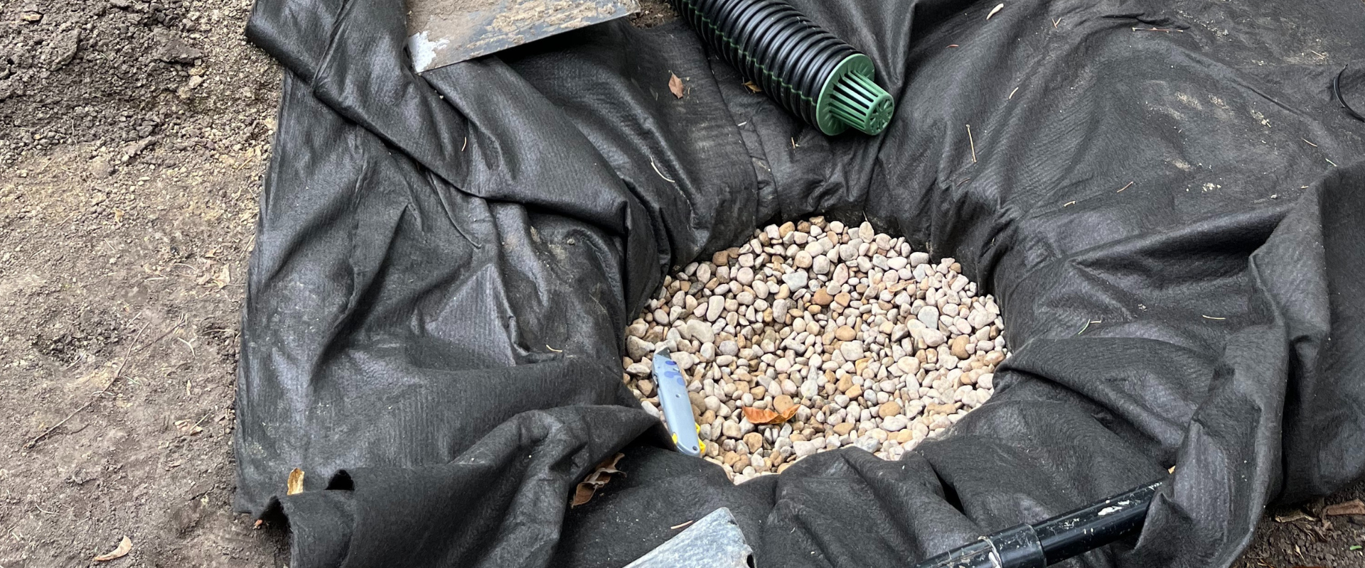 A black geomembrane liner filled with small gravel, surrounded by soil, with a green drainage pipe and a utility knife lying on it.
