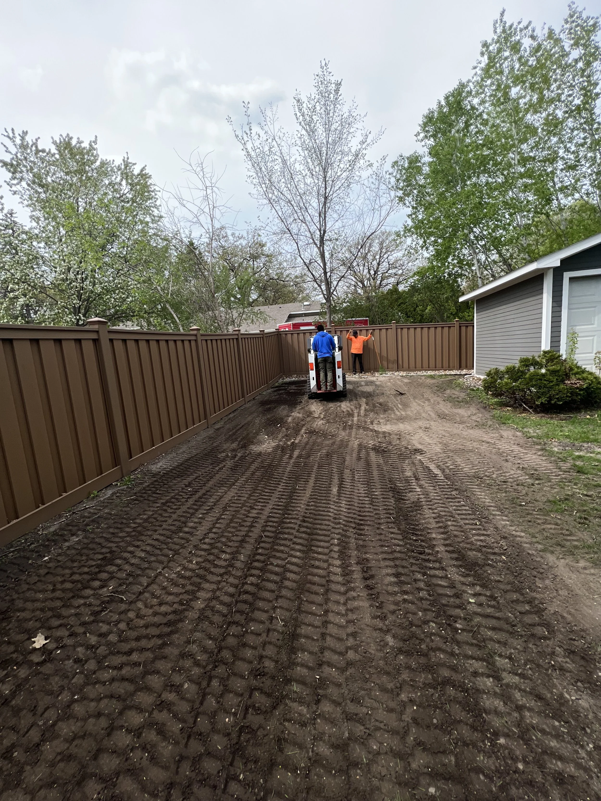 Two workers in safety gear spread fresh mulch along a newly leveled dirt driveway enclosed by a tall brown fence, with a house and trees in the background.