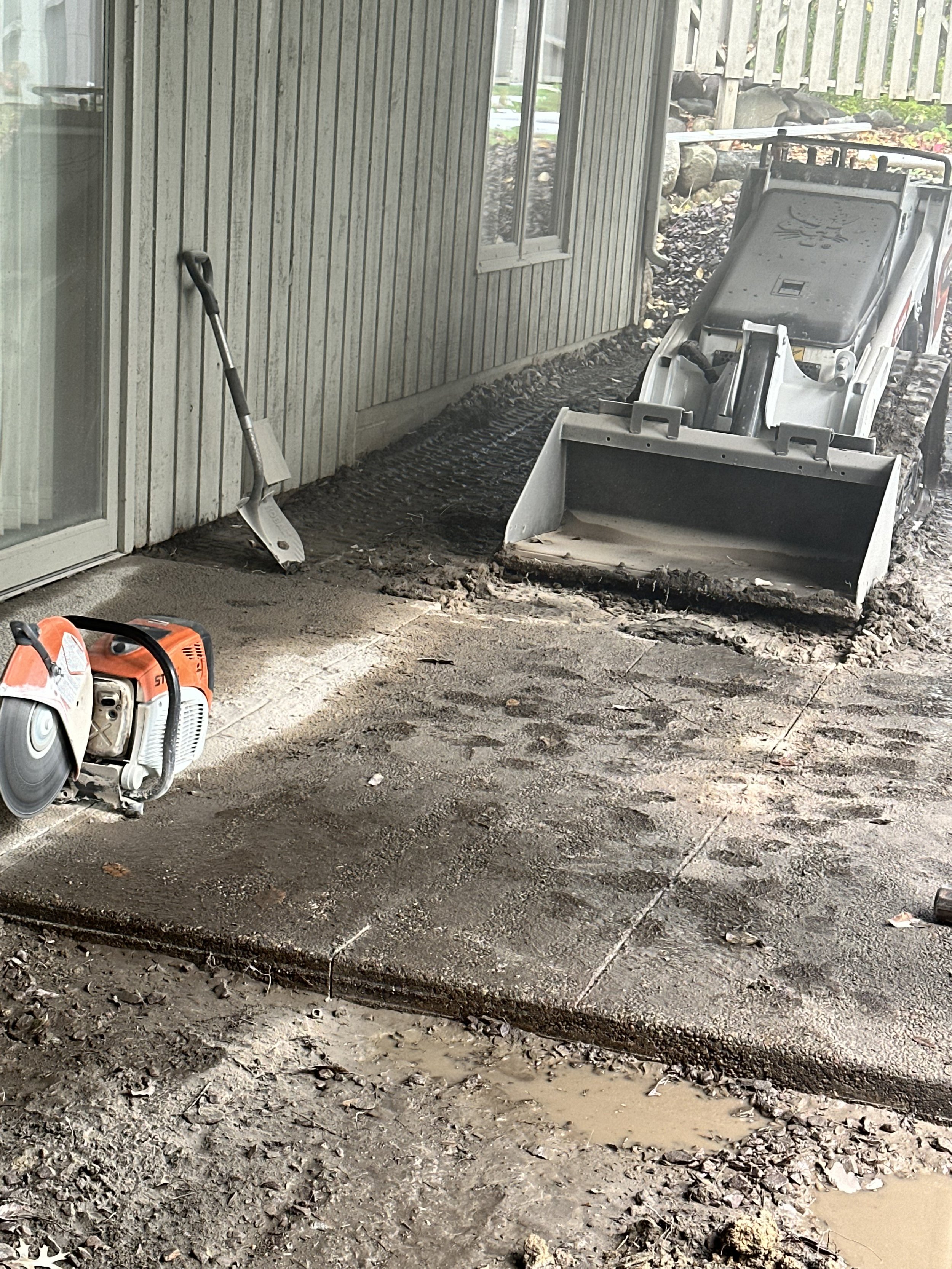 Construction site with a compacted dirt area, a saw machine, a shovel leaning against a wall, and a skid-steer loader with a bucket attachment next to a building with siding and a window.