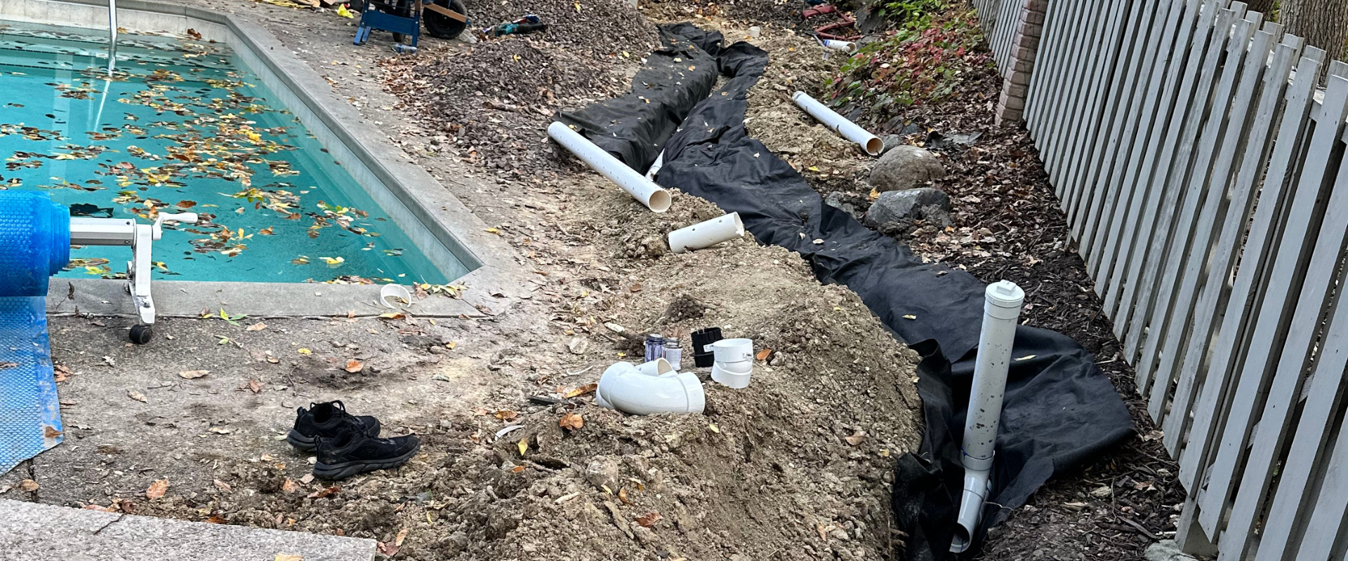 A backyard swimming pool under renovation with construction materials, including PVC pipes, black fabric liner, and dirt surrounding the pool.