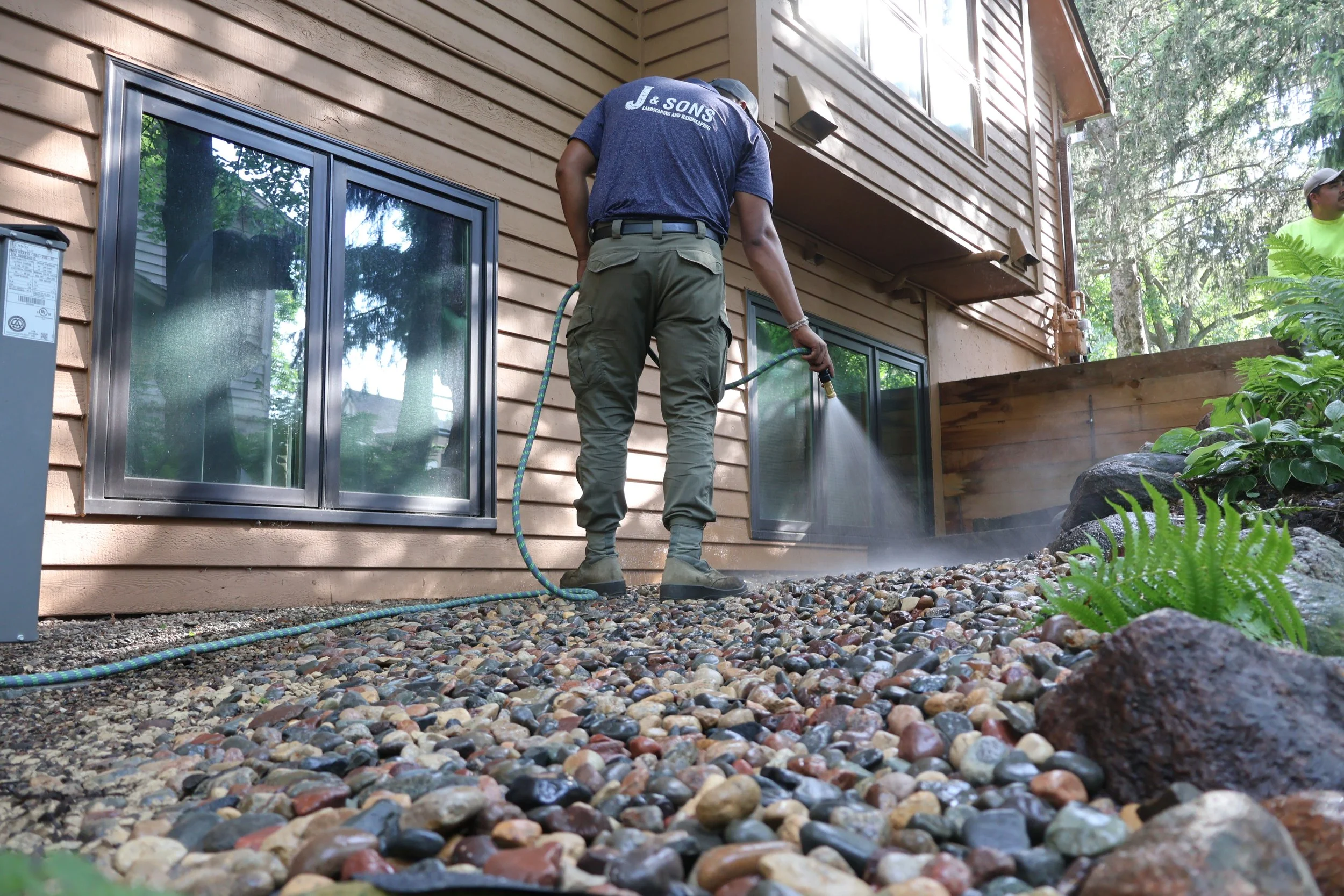 A person using a pressure washer to clean a gravel yard outside a house with wooden siding.
