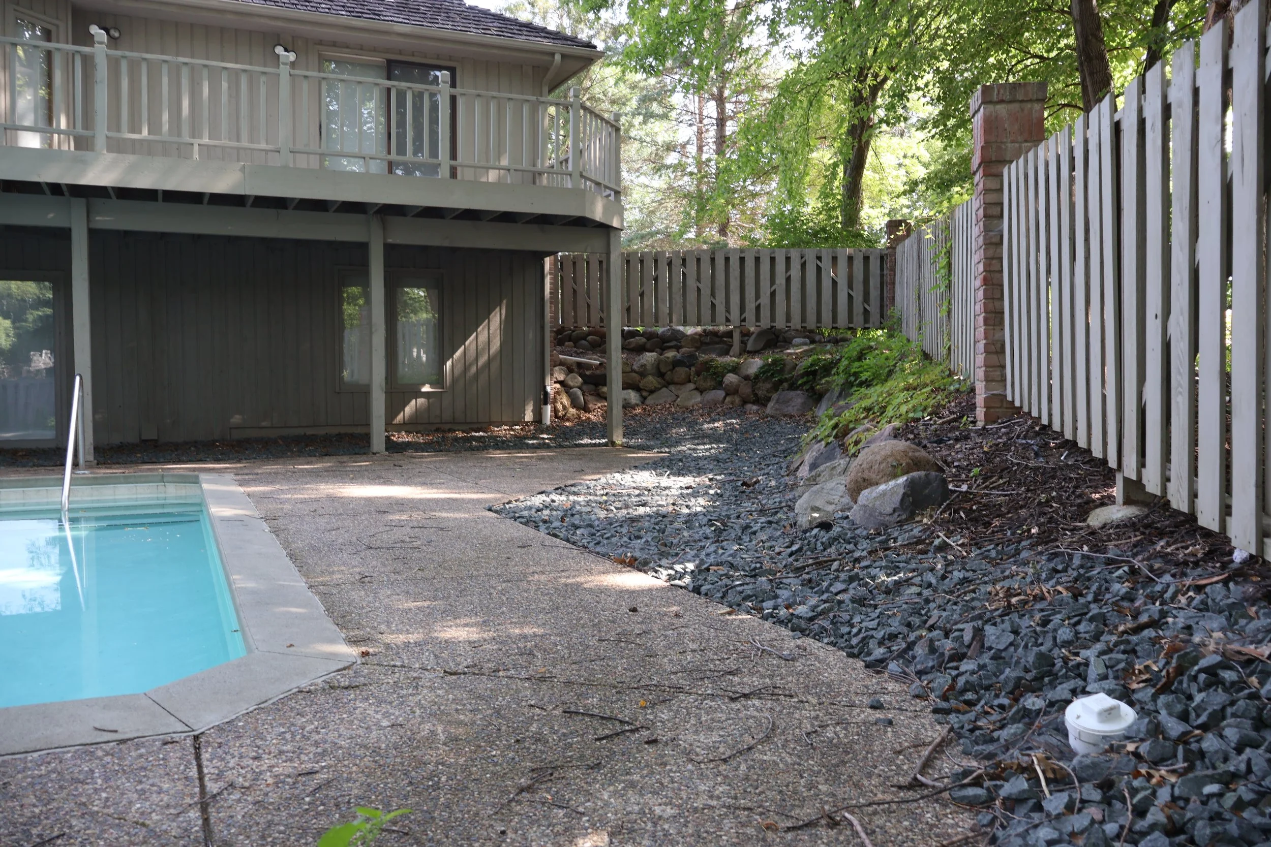 Backyard patio with kidney-shaped swimming pool, wooden deck, and a wooden fence, surrounded by trees and rocks.