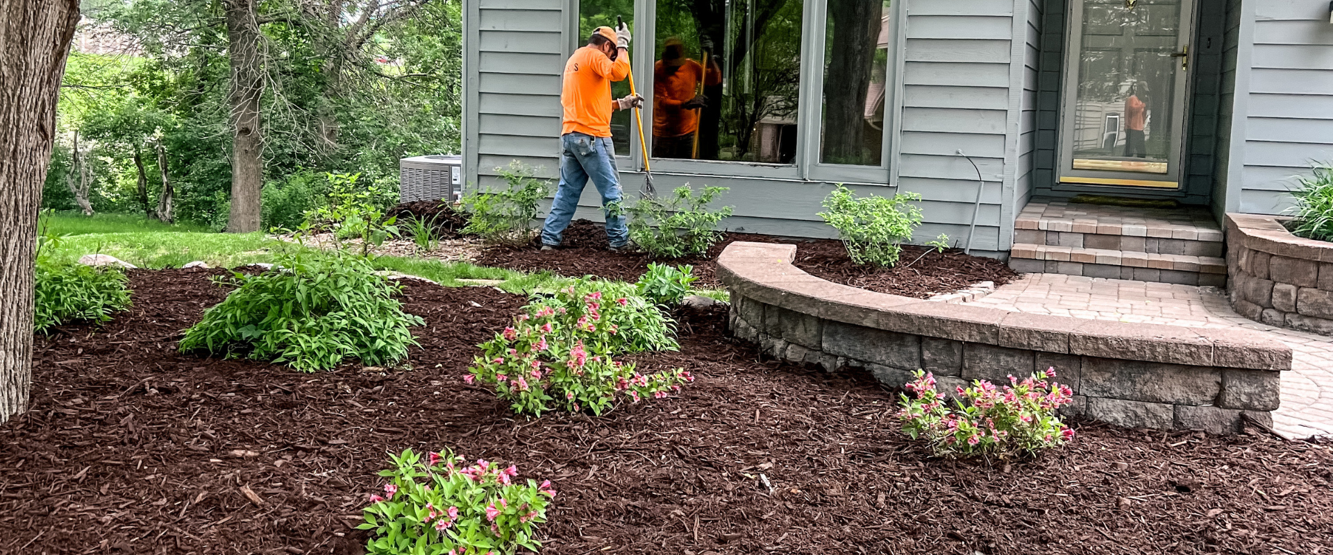 Person planting or gardening near a house with a landscaped yard, plants, mulch, and stone steps.