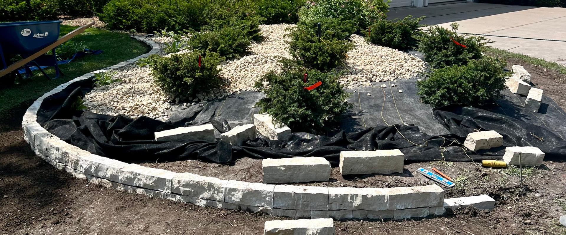 A garden bed under construction with a border of white stone bricks, black landscape fabric, and small shrubs planted. Construction tools and materials are visible.