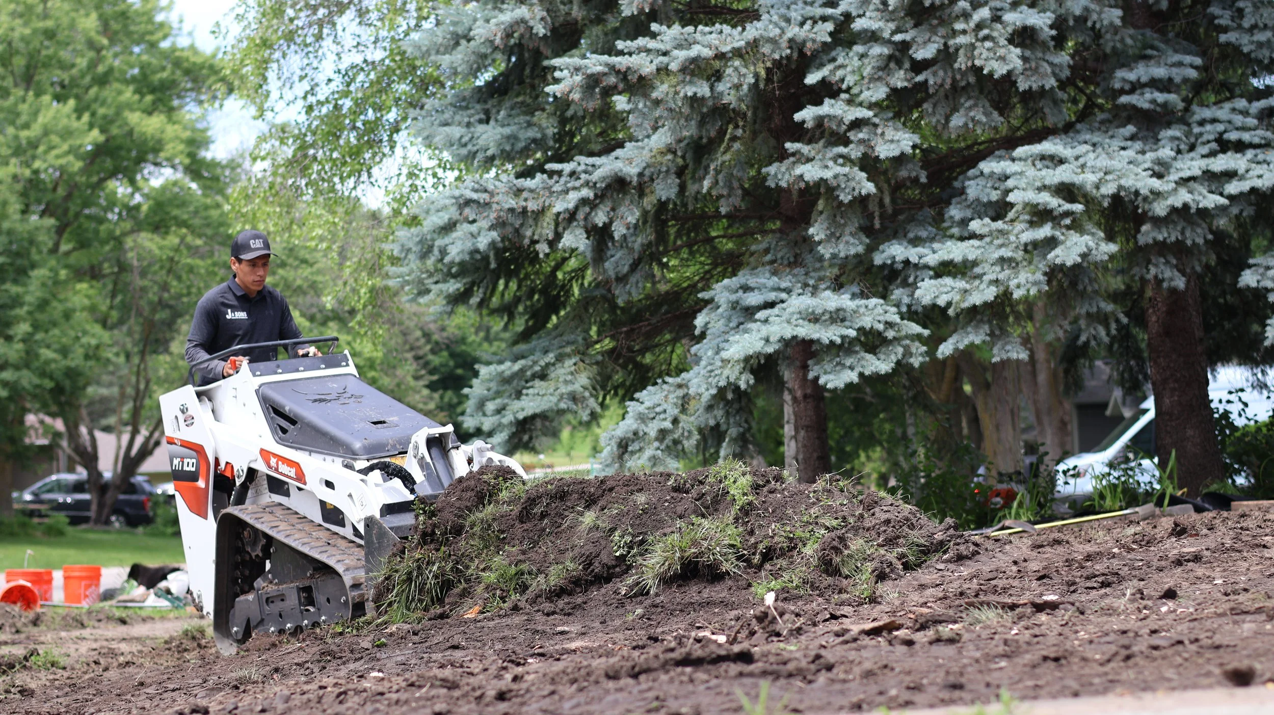 Man operating a compact track loader on a dirt construction site with trees and a house in the background.