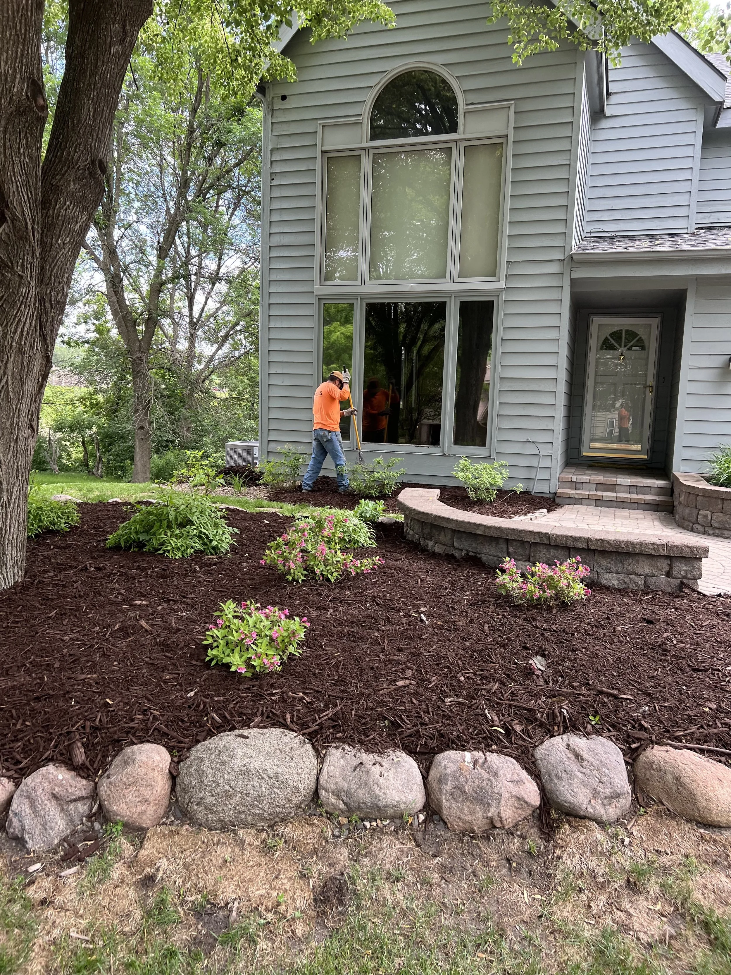 Man in blue jacket and black cap operating a red motorized lawn mower on brick patio next to a house, with a trailer filled with mulch and trees in the background.