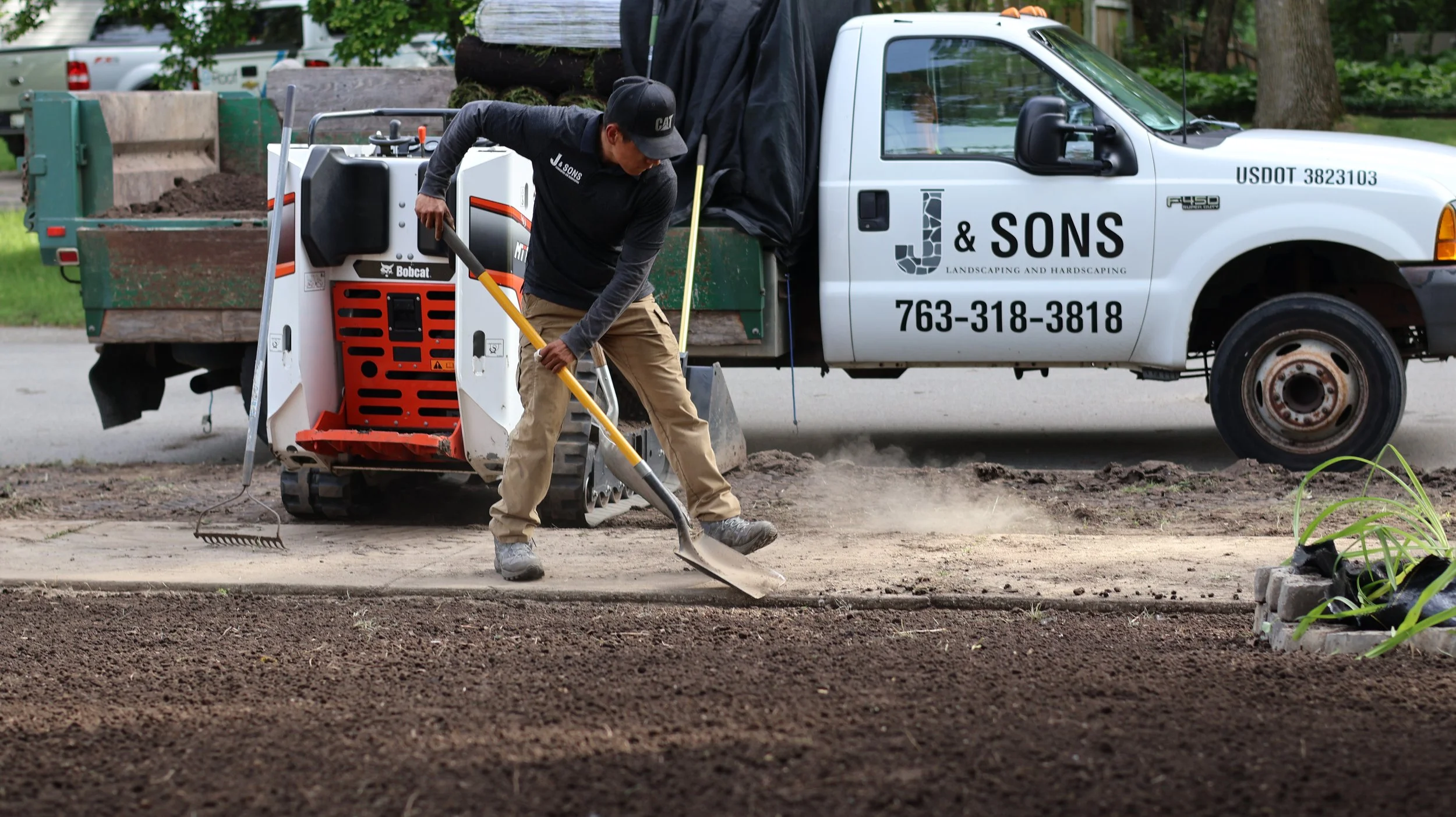 A man uses a shovel to level soil on a construction site with a compacted soil area in the foreground. Behind him, there is a truck labeled 'J & S Landscaping and Hardscaping' with a phone number, and a small Bobcat skid-steer loader.