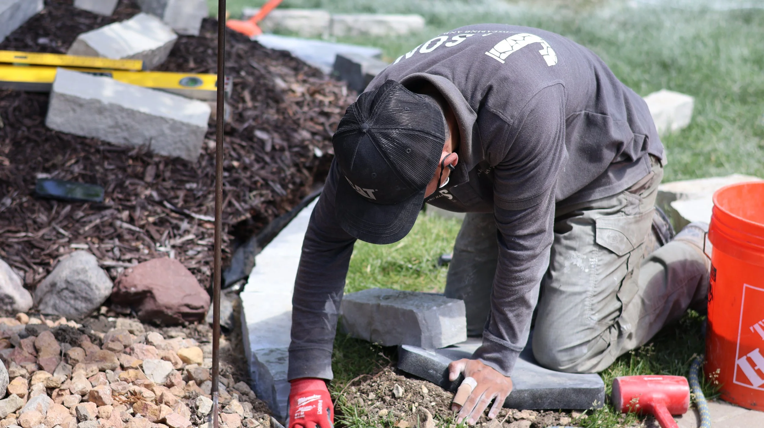 A worker kneels on the grass attaching stones to a flower bed border, wearing a black cap, grey long-sleeve shirts, and gloves, with tools and construction materials around him.