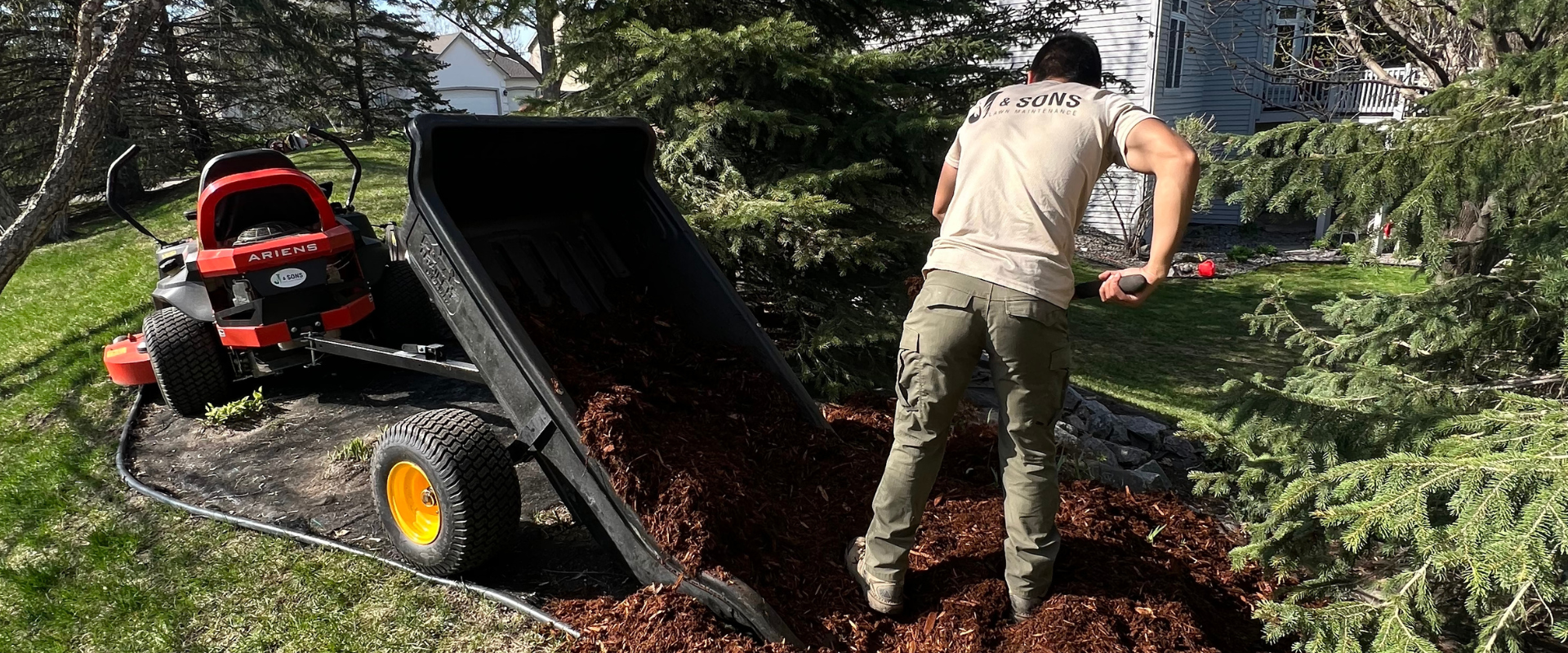 Man spreading mulch using a garden tractor on lawn in backyard with trees and house in background.