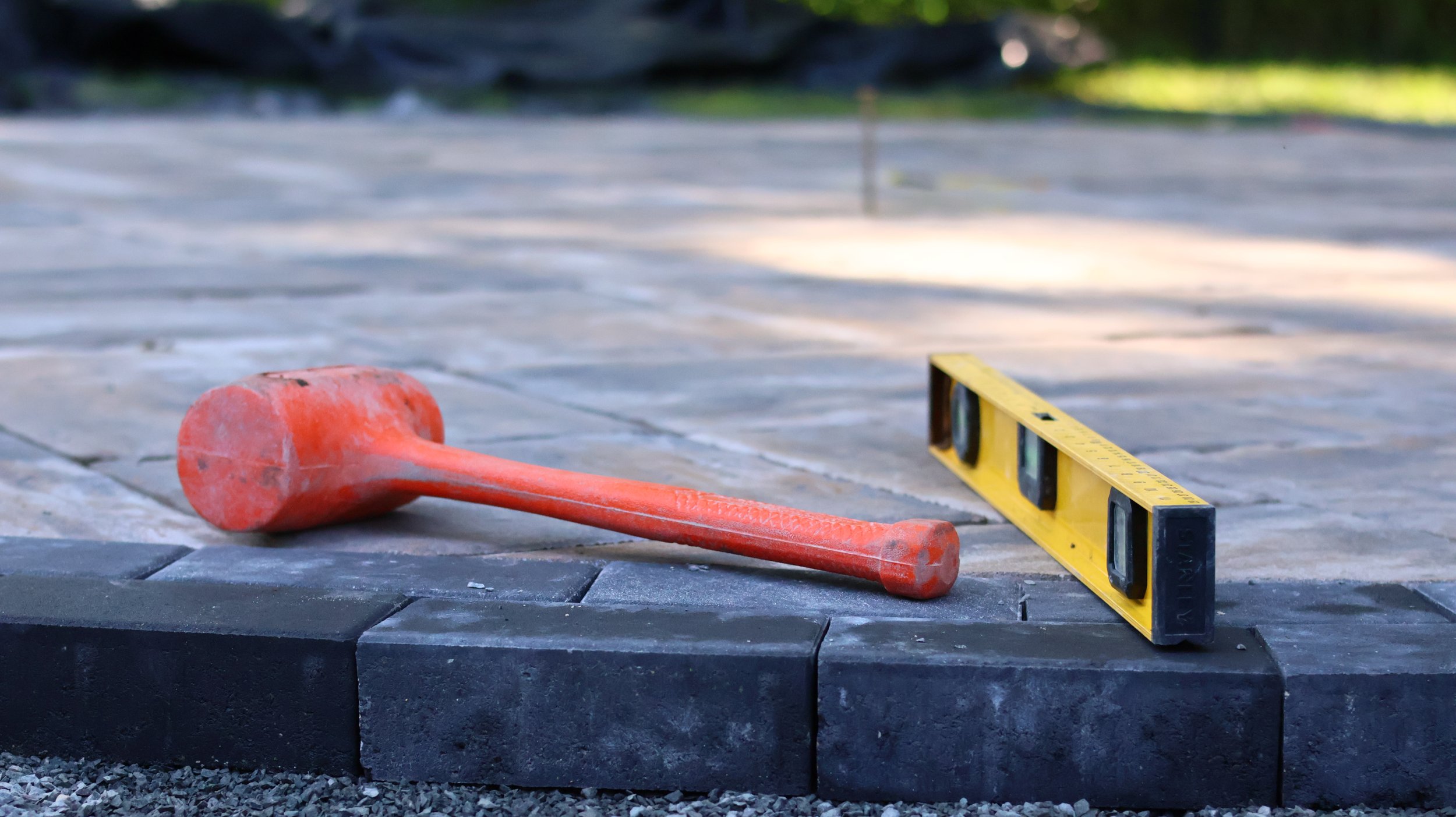 A rubber mallet and a yellow level tool placed on a newly laid stone paving surface outdoors during daylight.