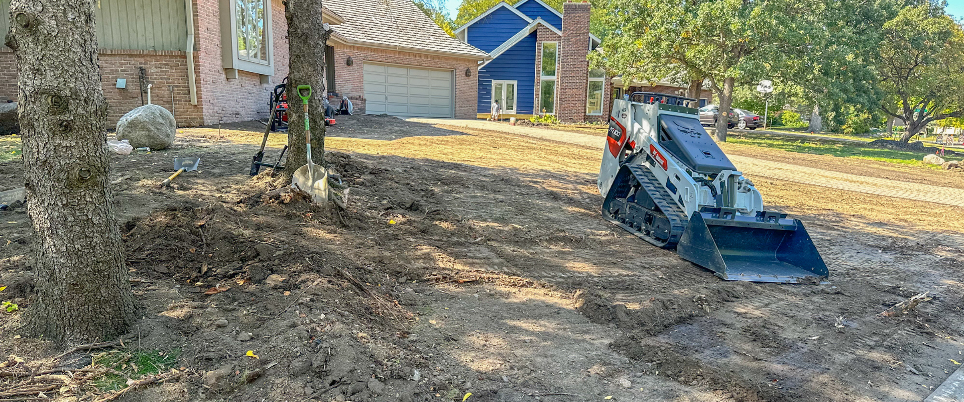 A residential yard under construction with dirt, trees, and a mini excavator. There's a house in the background with a blue house visible further back and several trees providing shade.