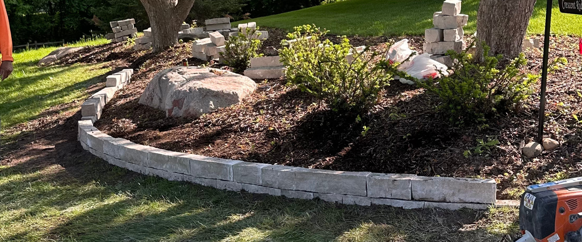 Garden bed being built with white bricks, containing soil, small bushes, and a large rock, with trees and a grassy lawn in the background.