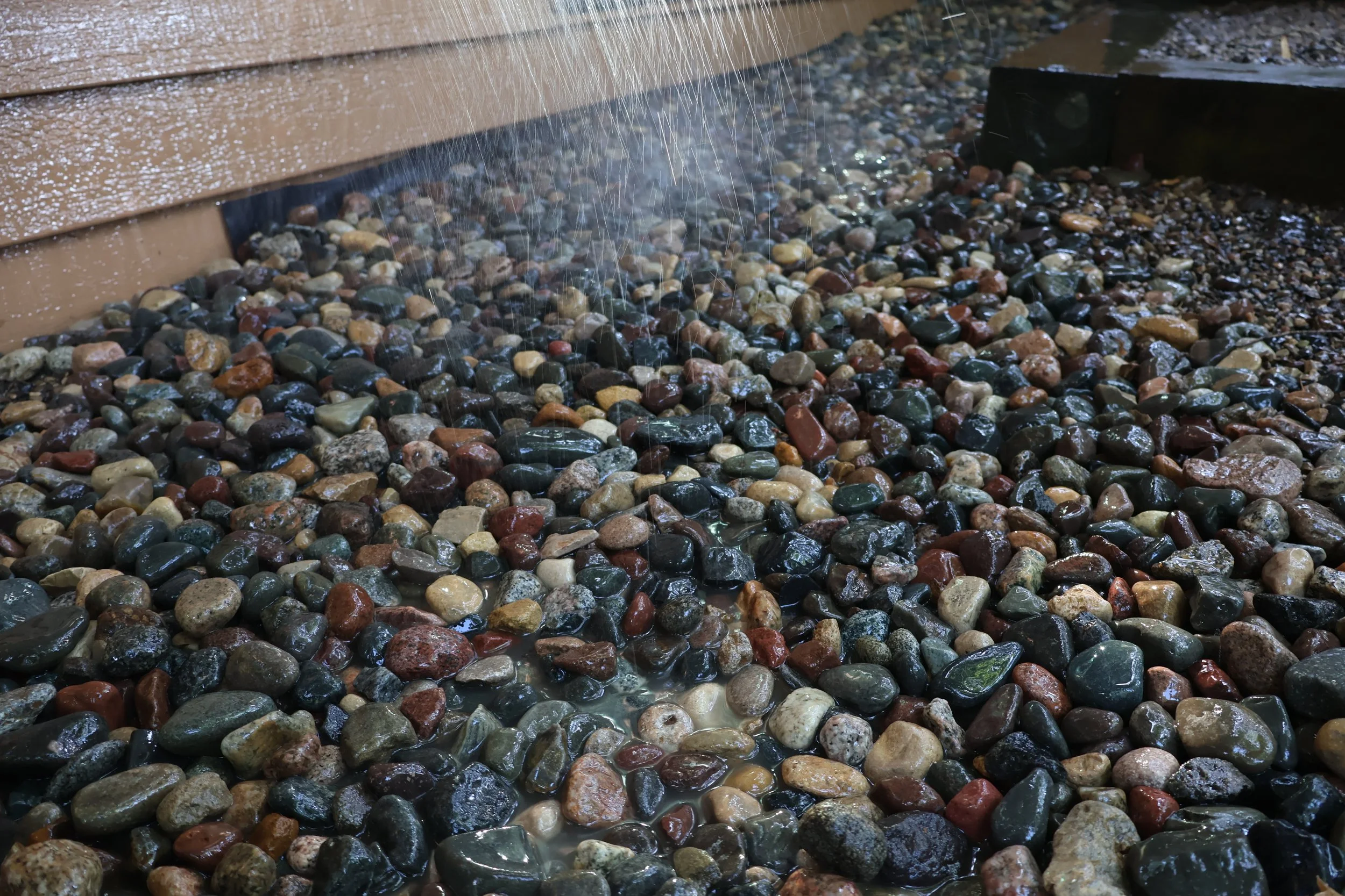 A close-up of a rainwater garden with colorful wet stones, a rain chain pouring water, and the ground decked with pebbles.