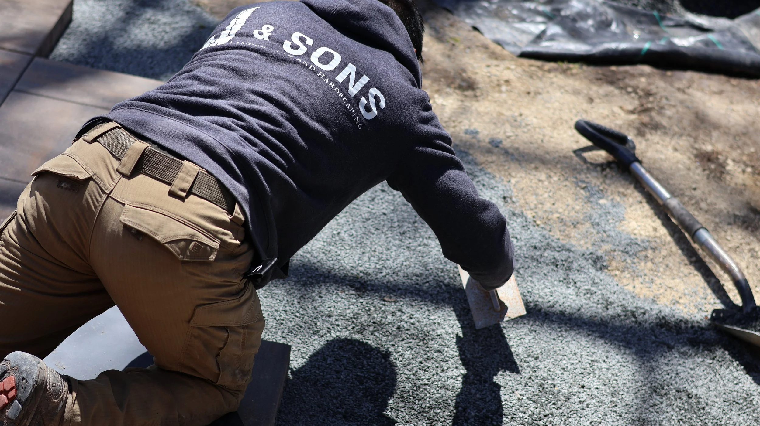 A construction worker is installing gravel on a sidewalk, kneeling on the ground with a trowel nearby.