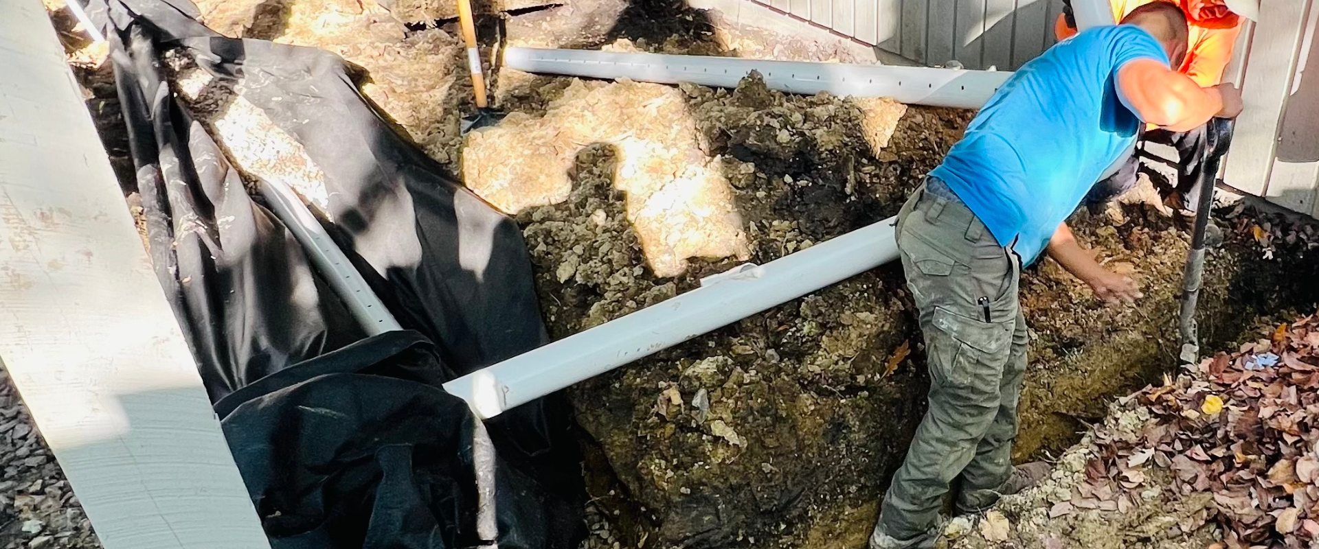 Construction worker installing or repairing a white gutter or drainage system outside a building, with black tarp and dirt around.