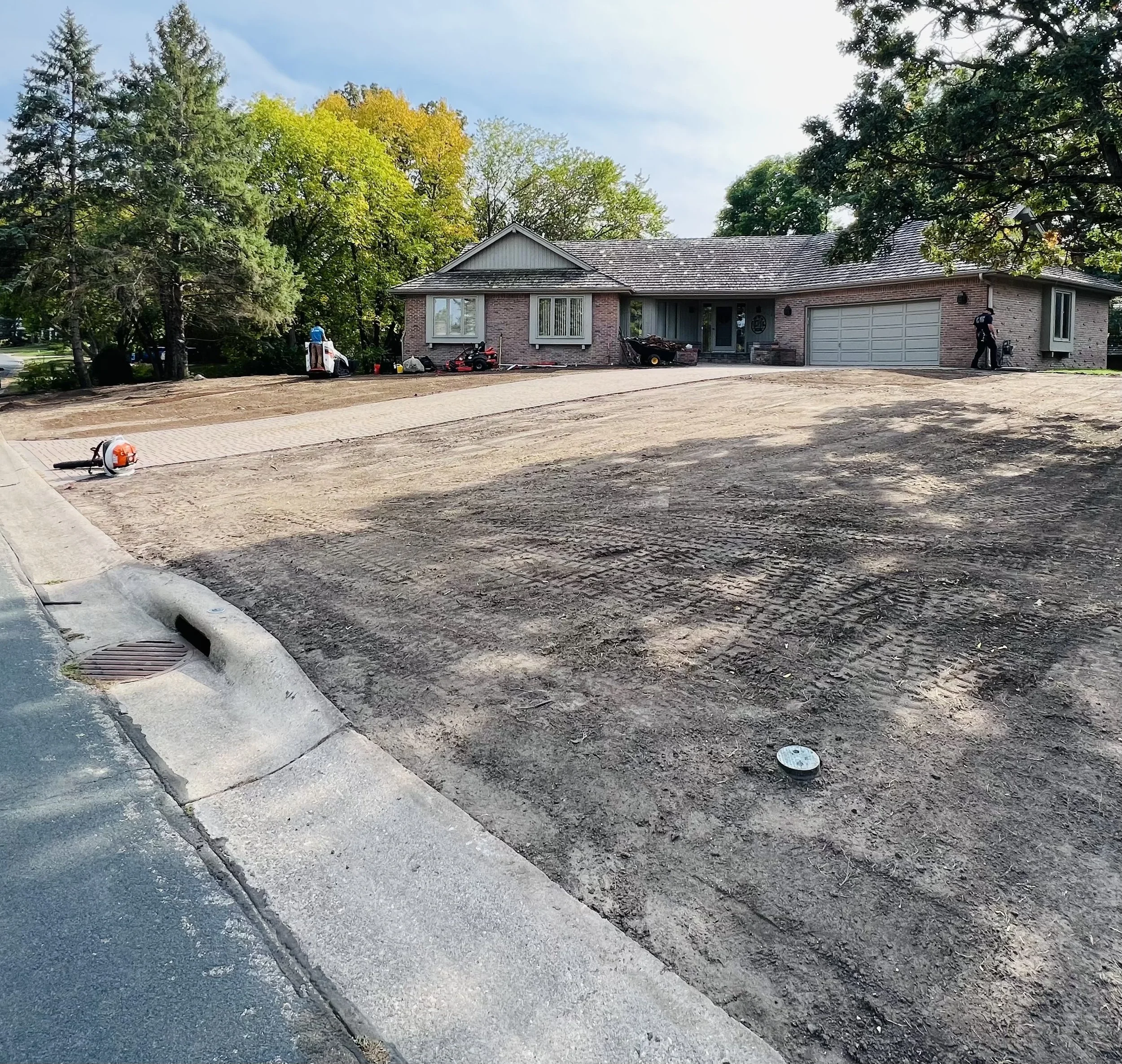 A house under construction with a dirt driveway, construction tools, and equipment visible in the yard, surrounded by trees with green and yellow leaves.