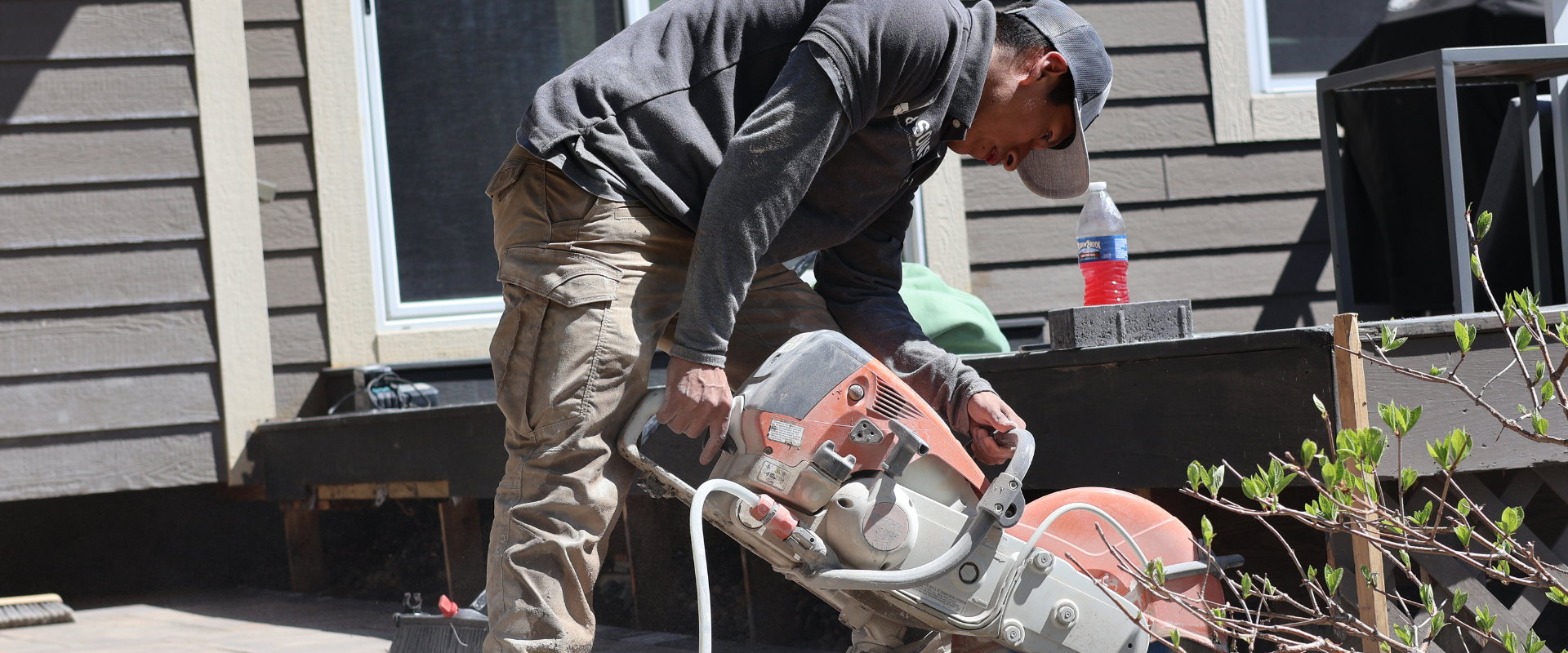 A man using a concrete cut-off saw to cut through a concrete surface outdoors on a porch, with building siding and windows in the background.