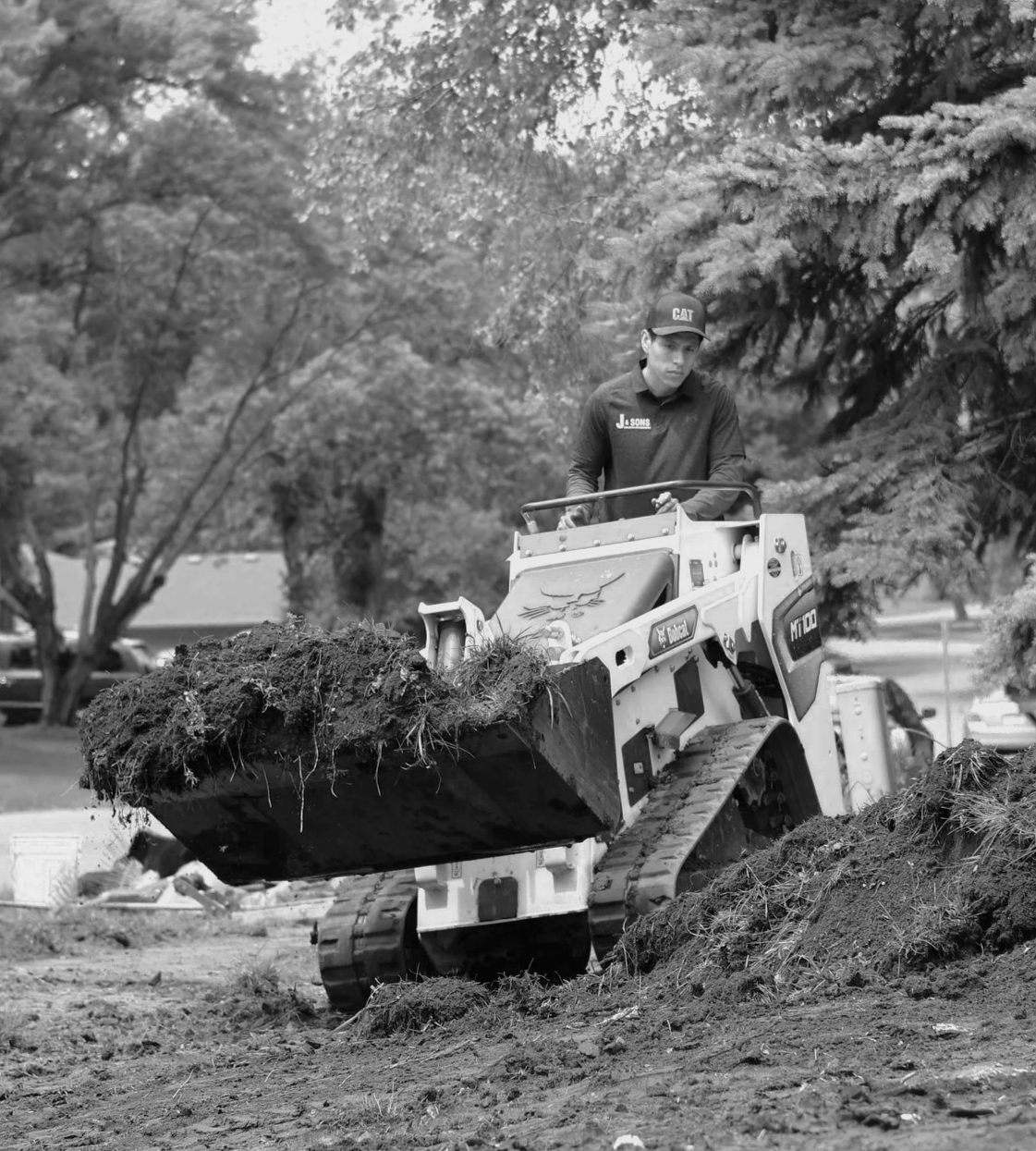 A man operating a small Bobcat construction vehicle moving dirt and roots in a landscaping or construction site. Trees and parked cars are visible in the background.