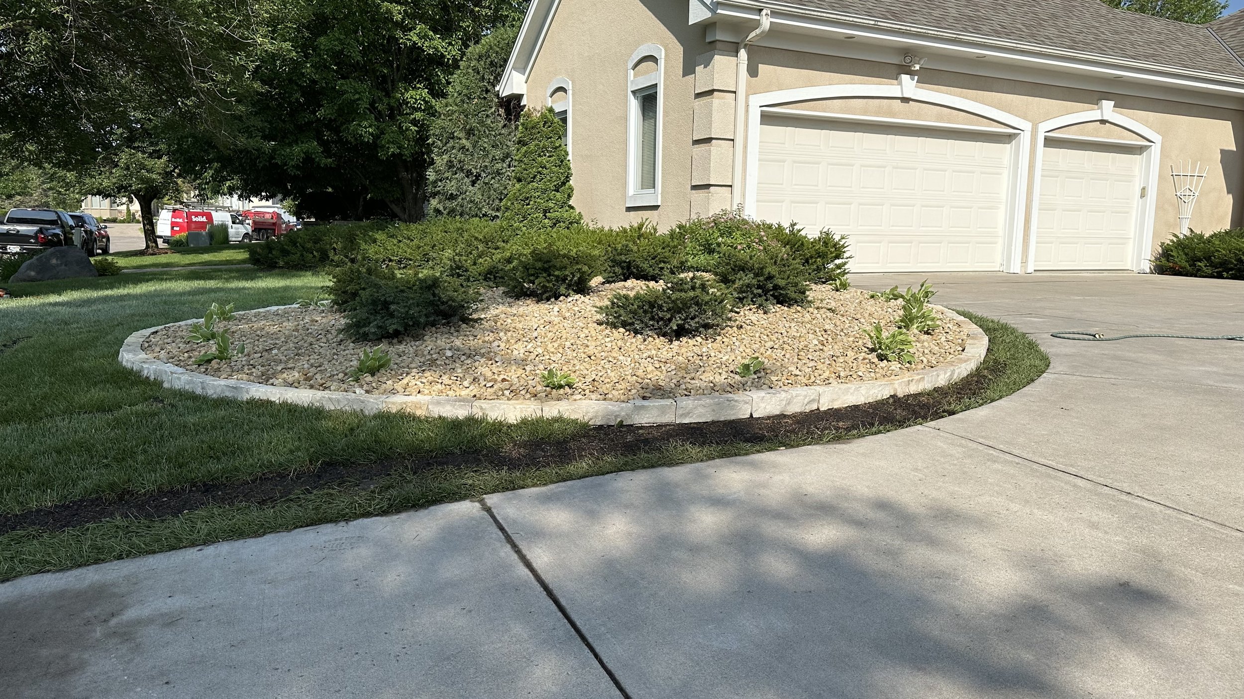 Residential front yard with a circular garden bed containing small shrubs and plants, edged with stone, next to a concrete driveway and house with a garage.
