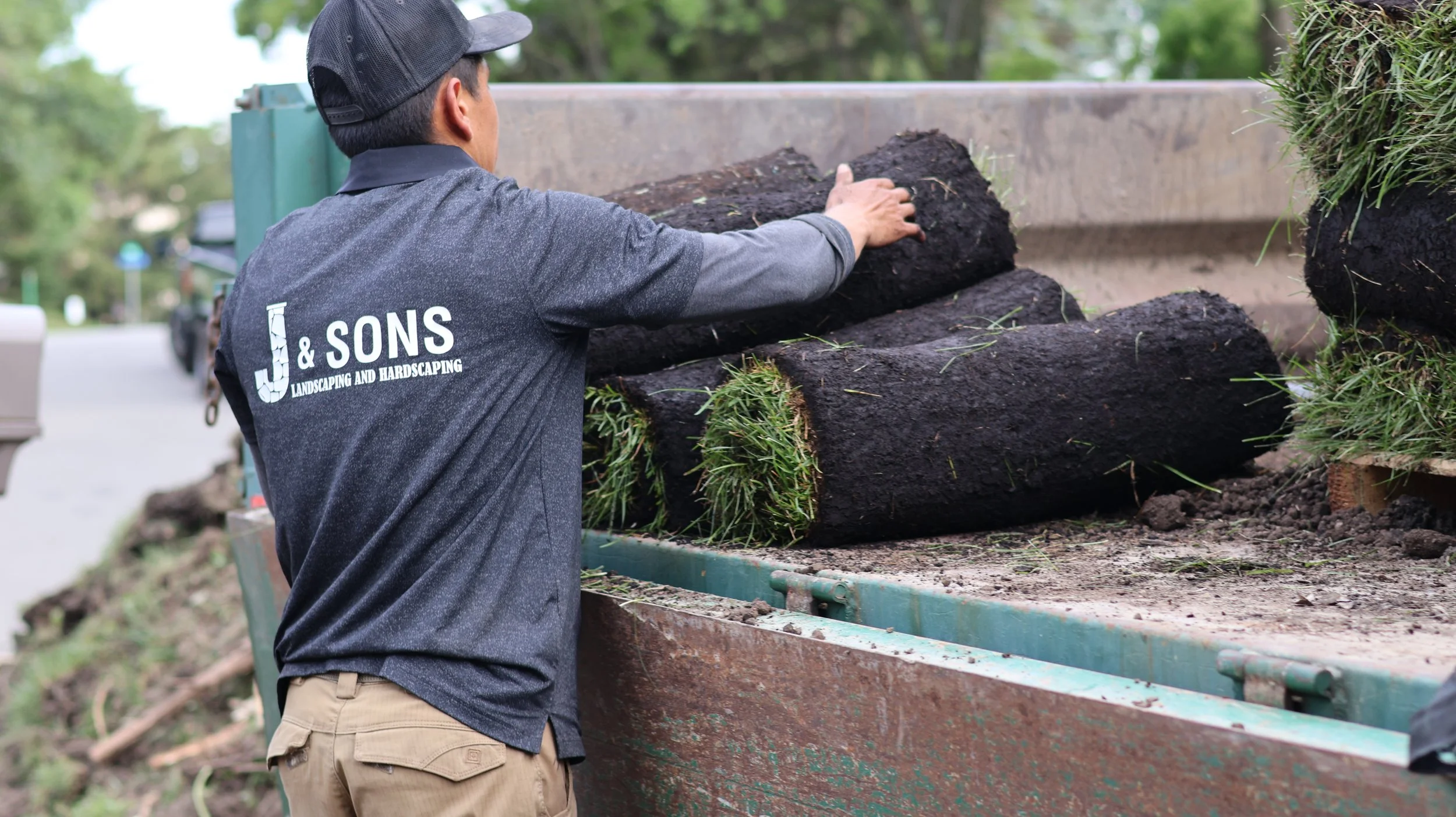 A worker loading or unloading large tree logs with grass and roots attached from a truck, wearing a dark uniform with 'J & Sons Landscaping and Hardscaping' on the back.