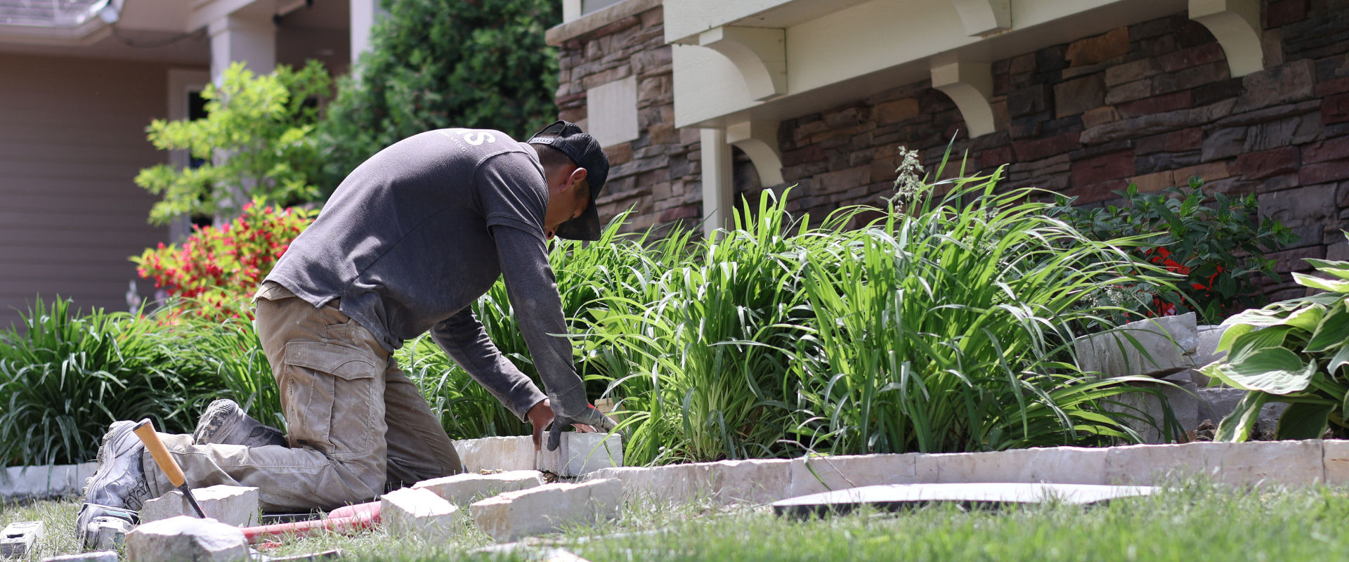 A man kneeling on the grass, laying bricks along the edge of a garden bed filled with green plants, in front of a house with a stone and siding exterior.
