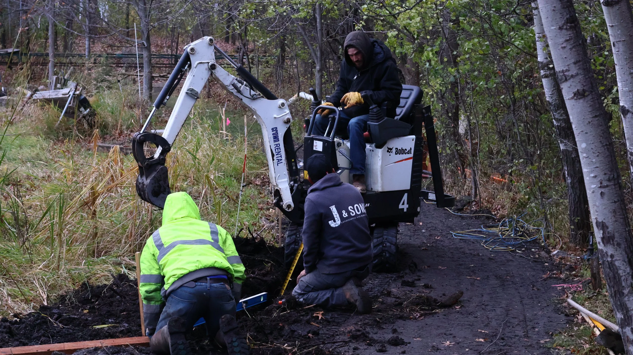 People working at a construction site in a wooded area, operating a small excavator marked 'Bobcat' and 'Crown Rental', with tools and wiring on the ground.