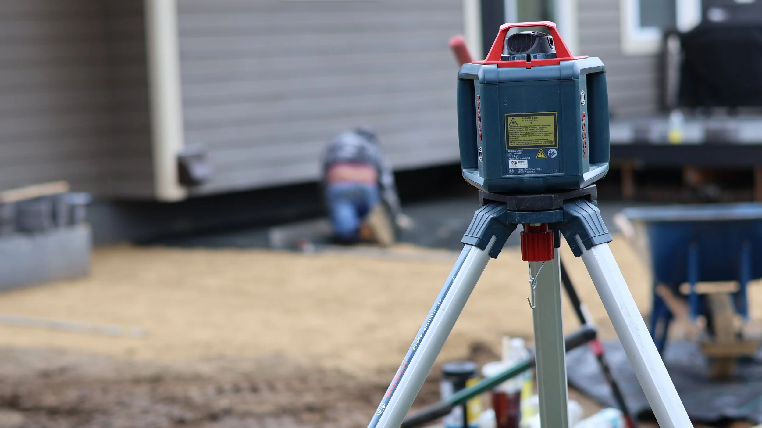 A construction laser level on a tripod at a construction site with a worker in the background.