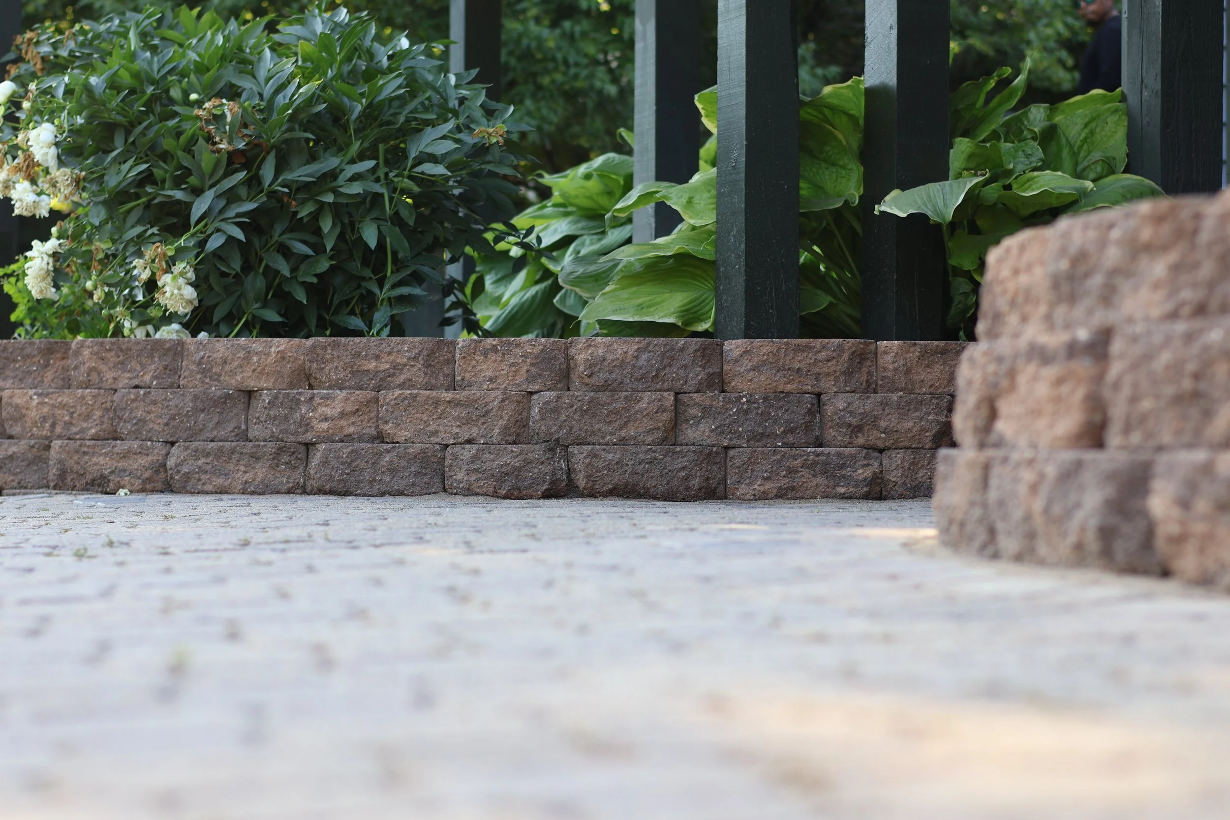 Close-up view of a brick garden wall with greenery and plants behind it, and a person partially visible in the background.