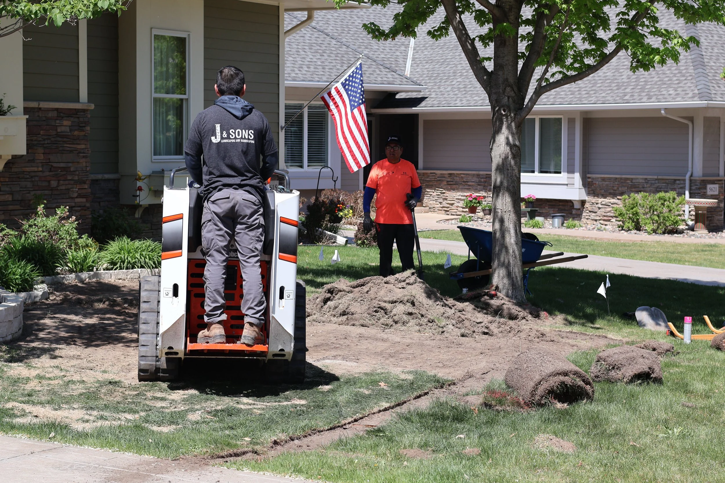 A person operating a small excavator in a residential yard during landscaping or gardening work, with two workers and a flag visible in the background.
