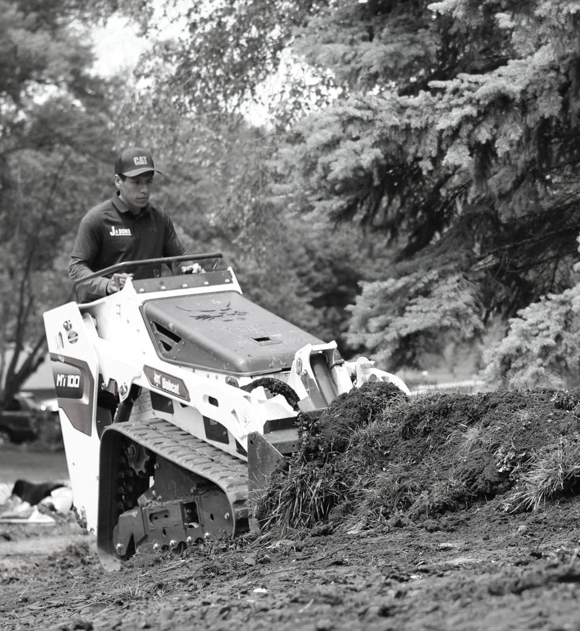 A person operating a Bobcat mini excavator working on a dirt mound outdoors, with trees in the background.