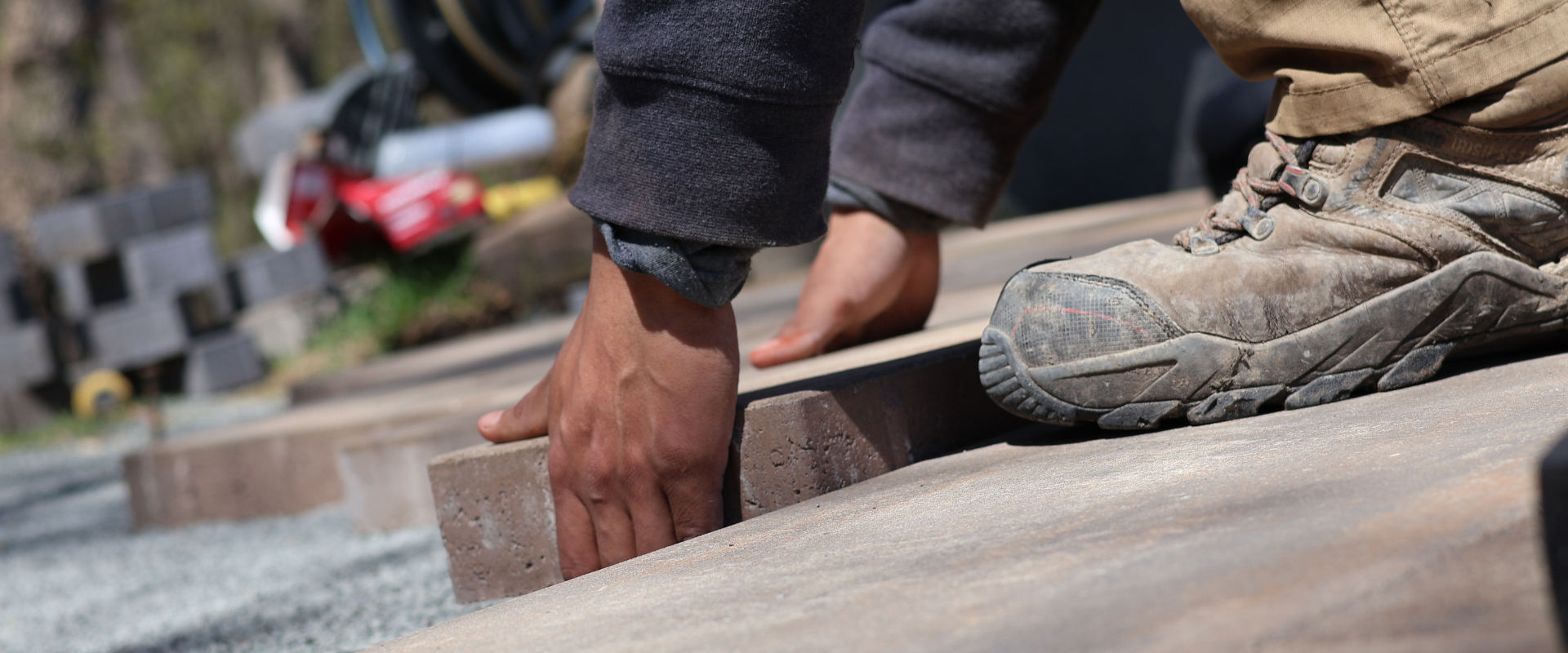 A worker in dusty work boots and long pants installing bricks on a pathway, using his hands to place a brick in position.