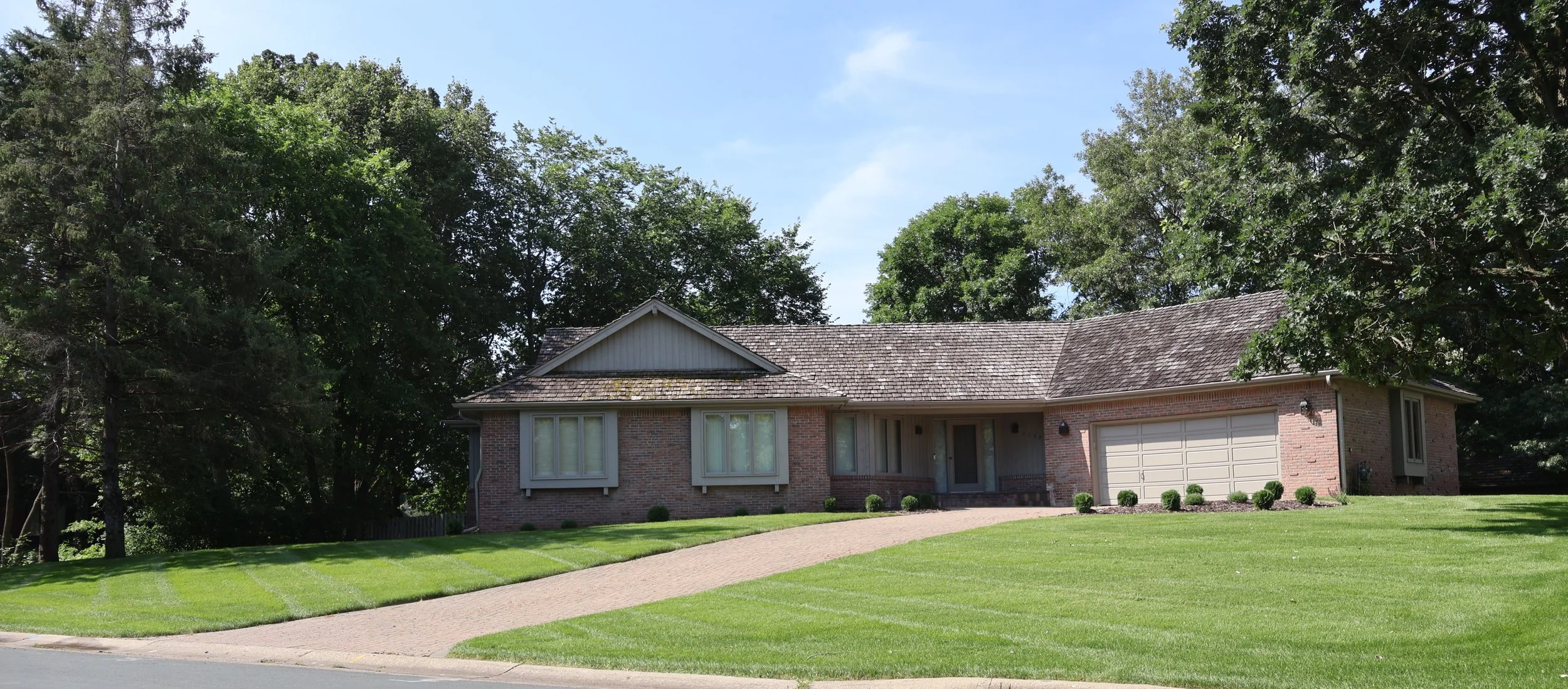 A single-story brick house with a gable roof, surrounded by a well-maintained lawn and tall trees in the background.