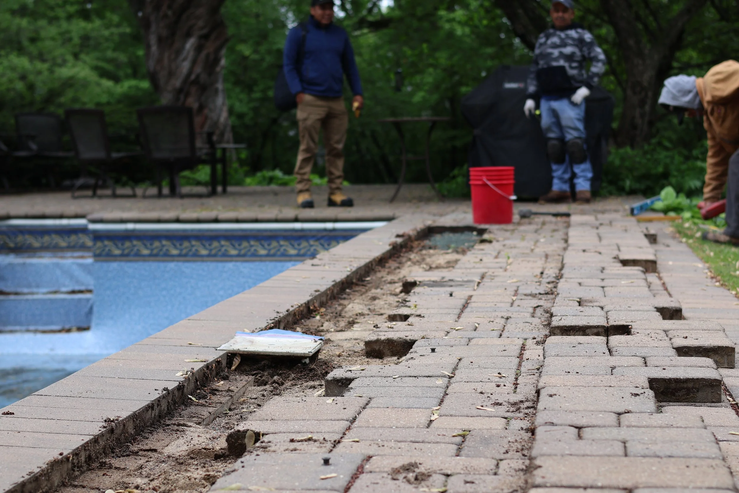 People working on installing or repairing a brick sidewalk beside a swimming pool outdoors, with trees and outdoor furniture in the background.