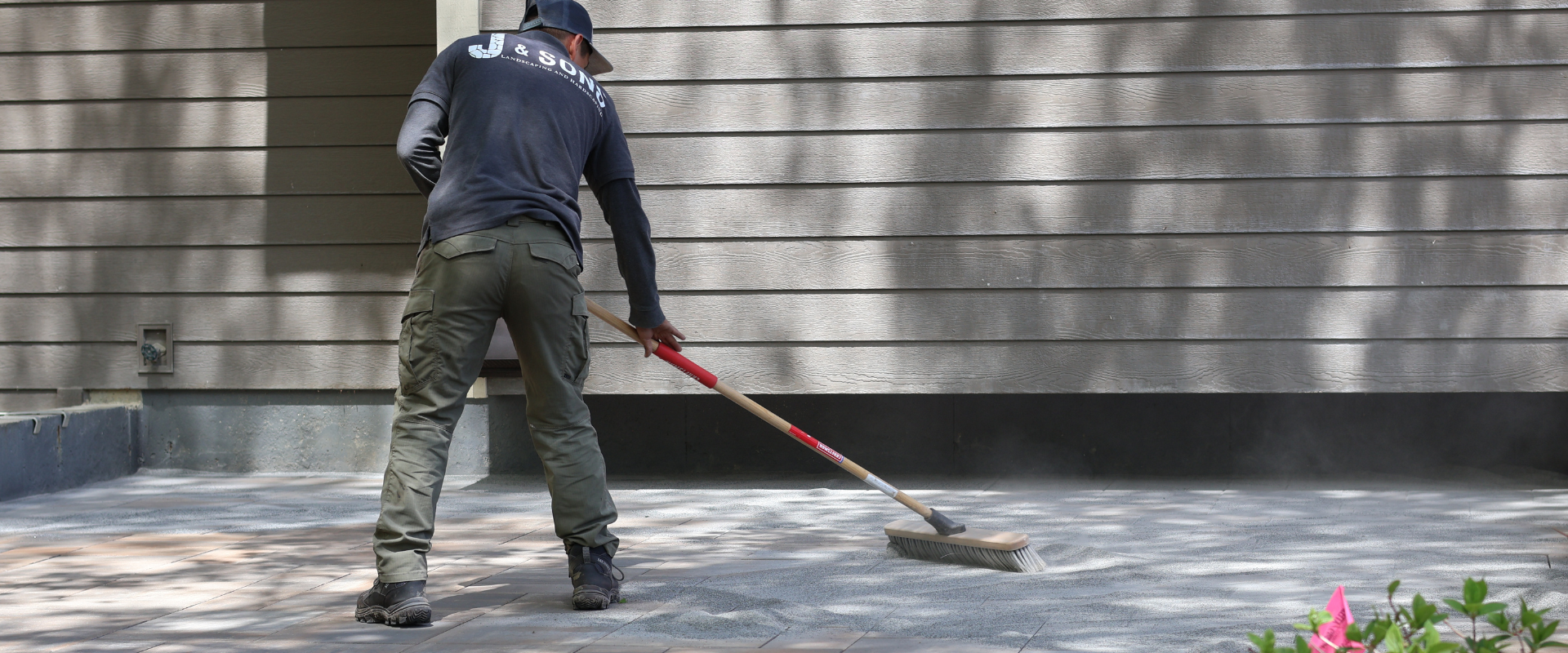 A man sweeping a patio outdoors near a wood-paneled house wall.