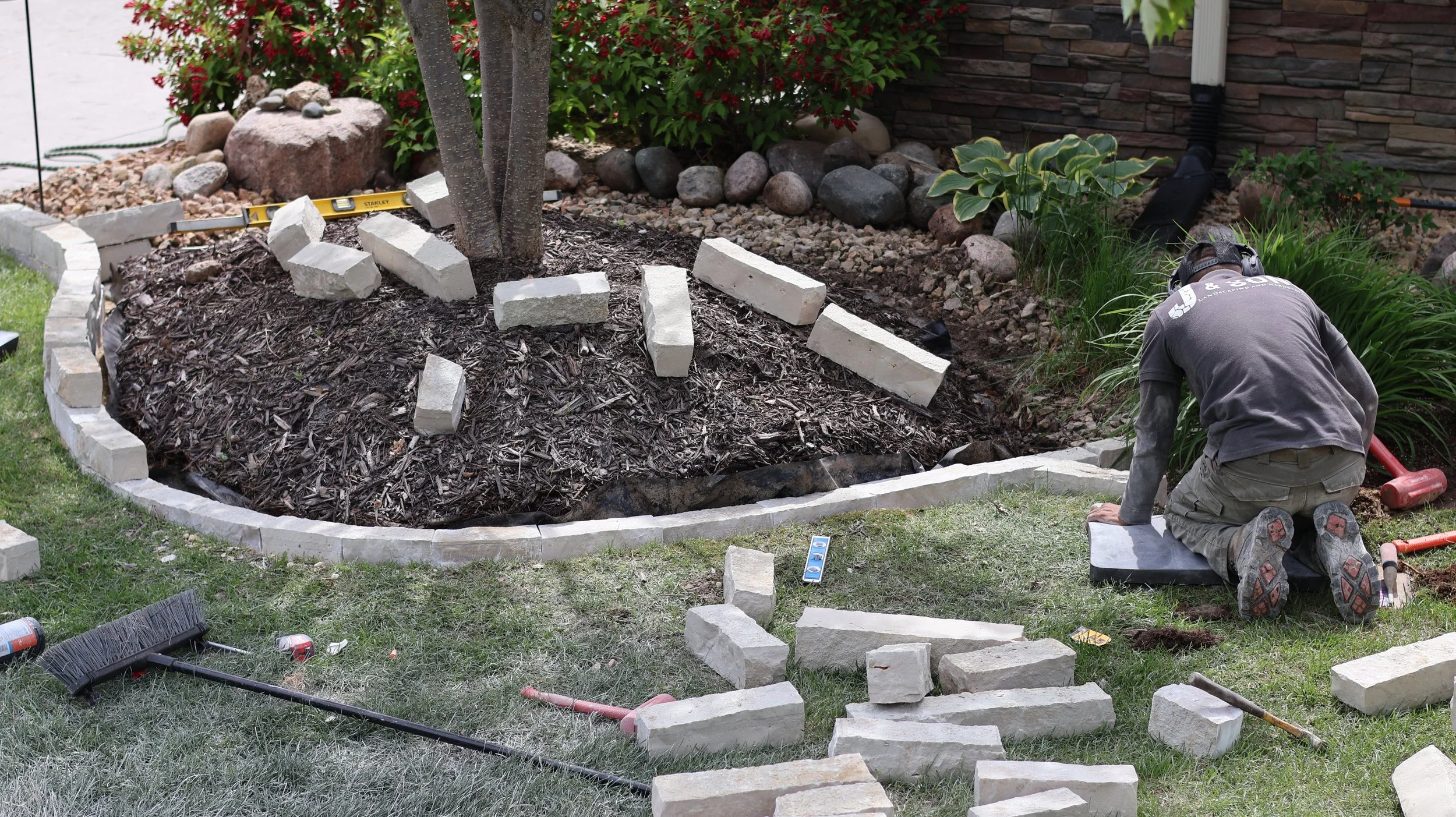 A person kneeling on the ground installing stone edging around a garden bed with mulch, bricks, and garden tools nearby.