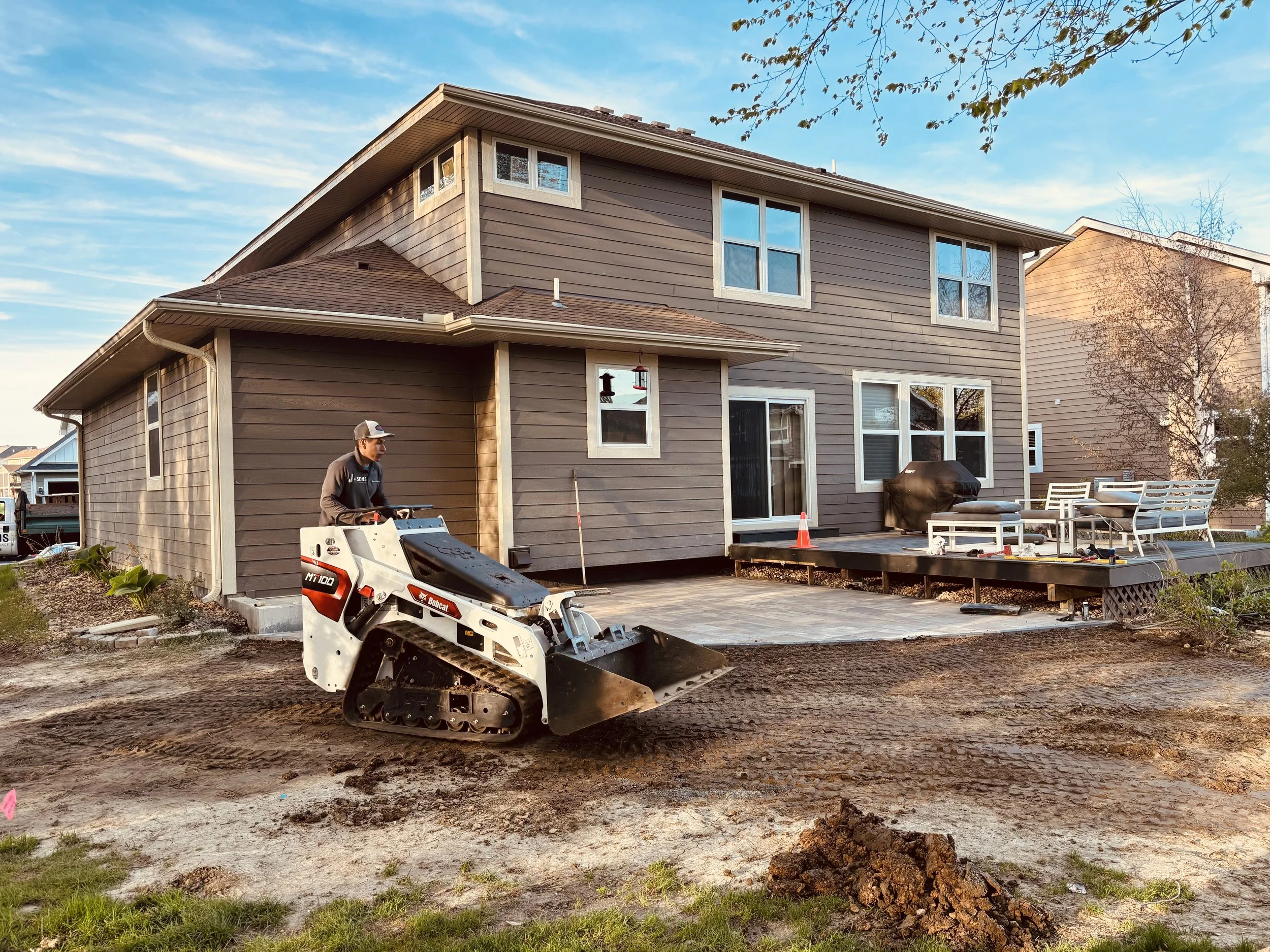 A house with brown siding and multiple windows in a suburban neighborhood. The yard is under construction with dirt and a small patch of grass. A person in work clothes is operating a mini bulldozer. Outdoor furniture, including a table and chairs, is visible on the patio, and there are trees and a partly cloudy sky in the background.