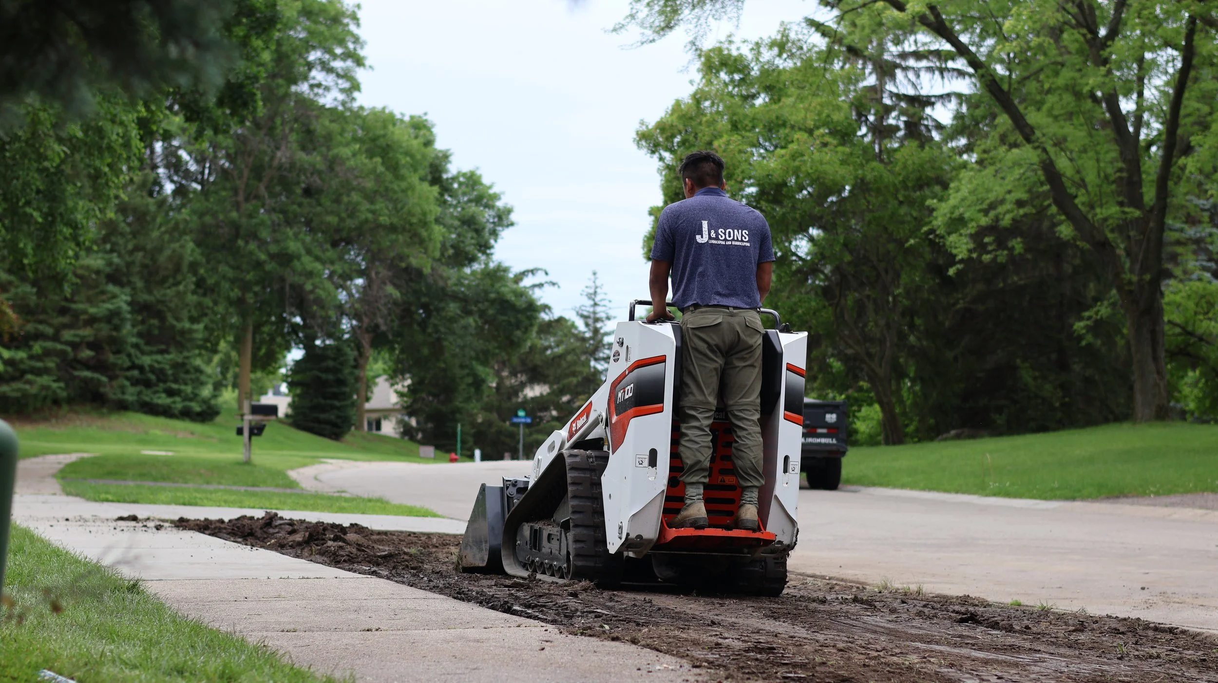 A worker operating a small tracked skid steer with a bucket attachment, grading or leveling a newly paved sidewalk in a residential neighborhood with trees and houses in the background.