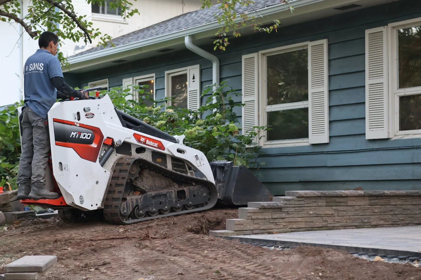 A person operating a small tracked skid steer loader on a patch of dirt beside a white house with a beige roof and trees in the background.