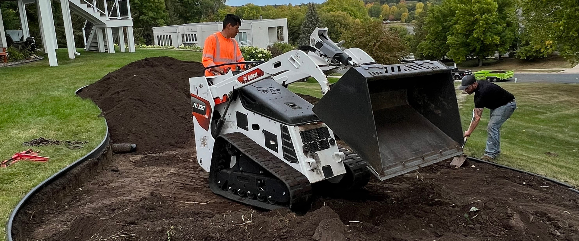 Two workers are using a compact Bobcat loader to level soil on a landscaped yard, with one pushing soil and the other digging with a shovel.