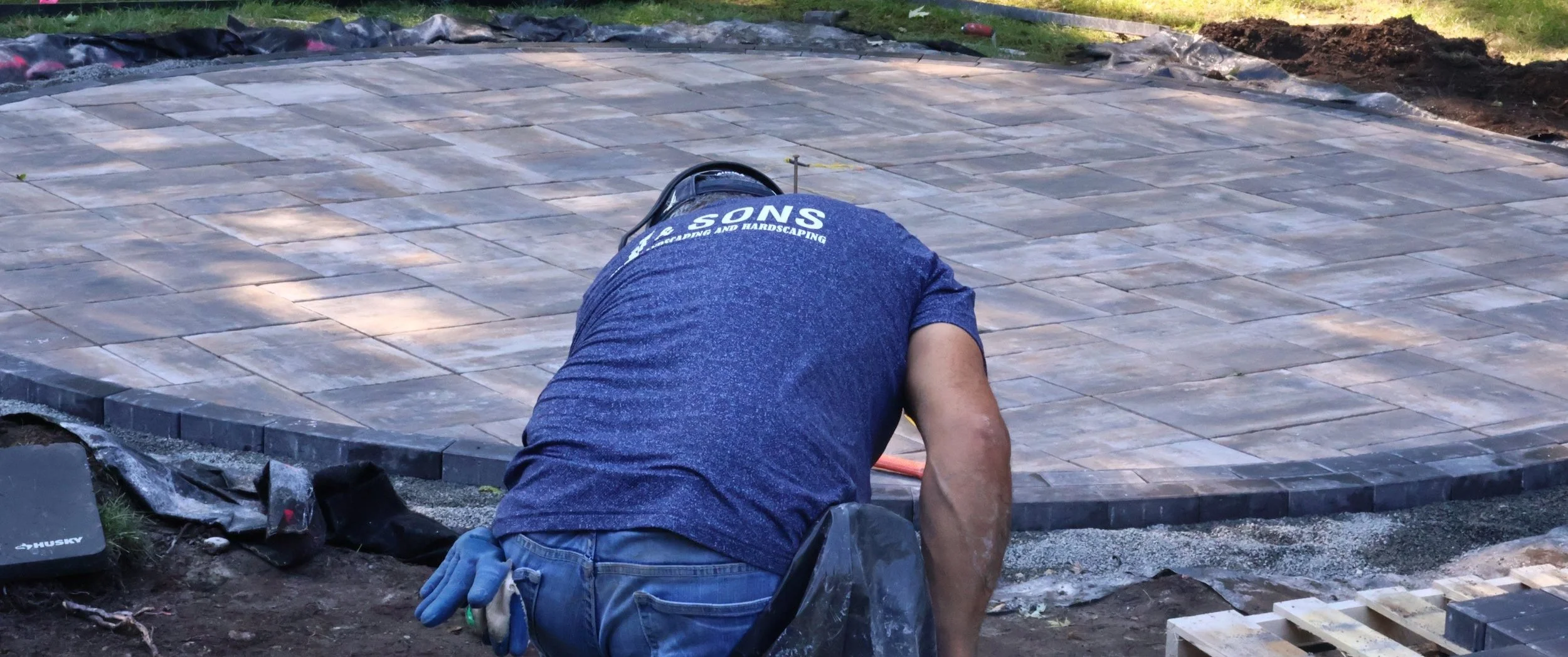 A worker installing stone pavers on a patio, wearing a blue shirt with white text, kneeling on the ground, with tools and construction materials nearby.