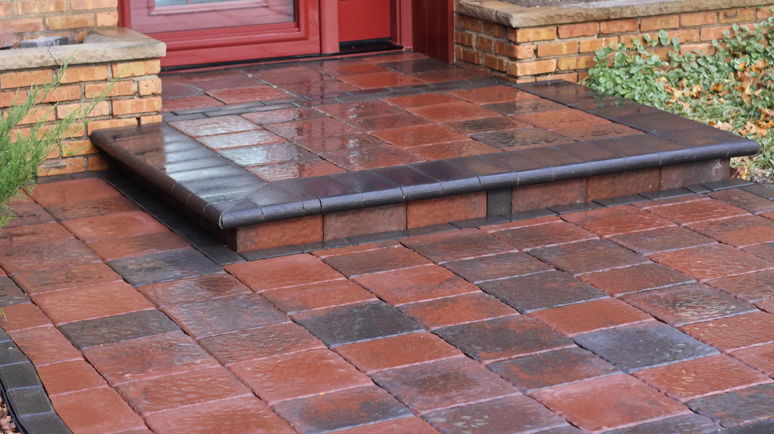 Wet brick front porch with a red door, bordered by dark tile trim and surrounded by green plants.