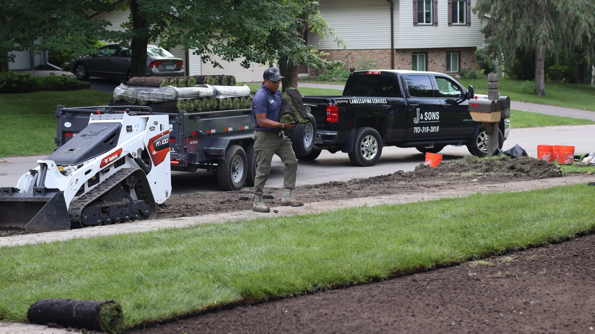 A landscaper in a blue shirt and khaki pants working on a lawn renovation, holding a sod roll, with a trailer full of sod, a small excavator, and a black landscaping truck in a residential neighborhood with trees and houses.