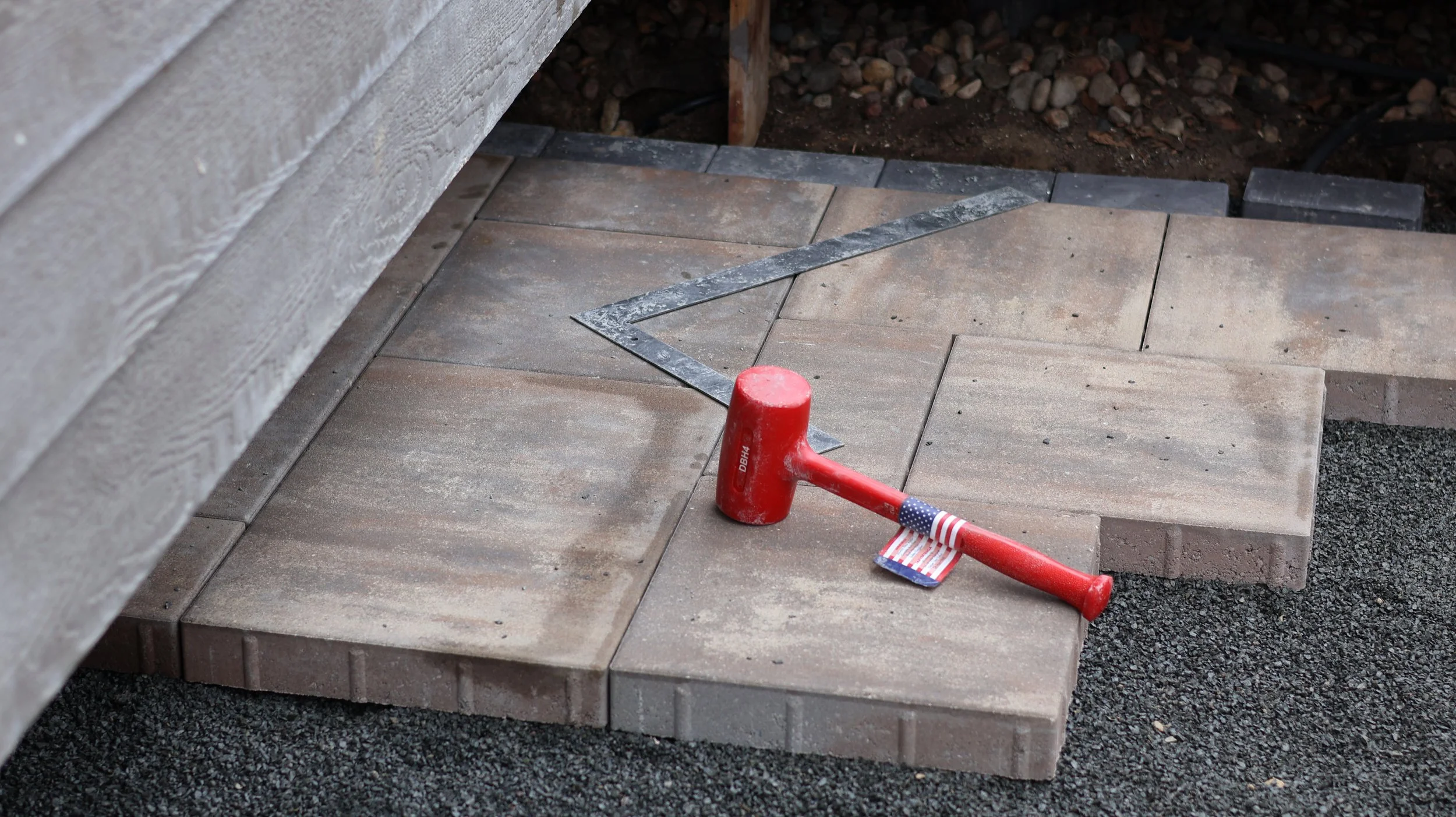 A construction site with a partially completed paver patio, a rubber mallet with an American flag design on the handle, and a metal square tool on the bricks.