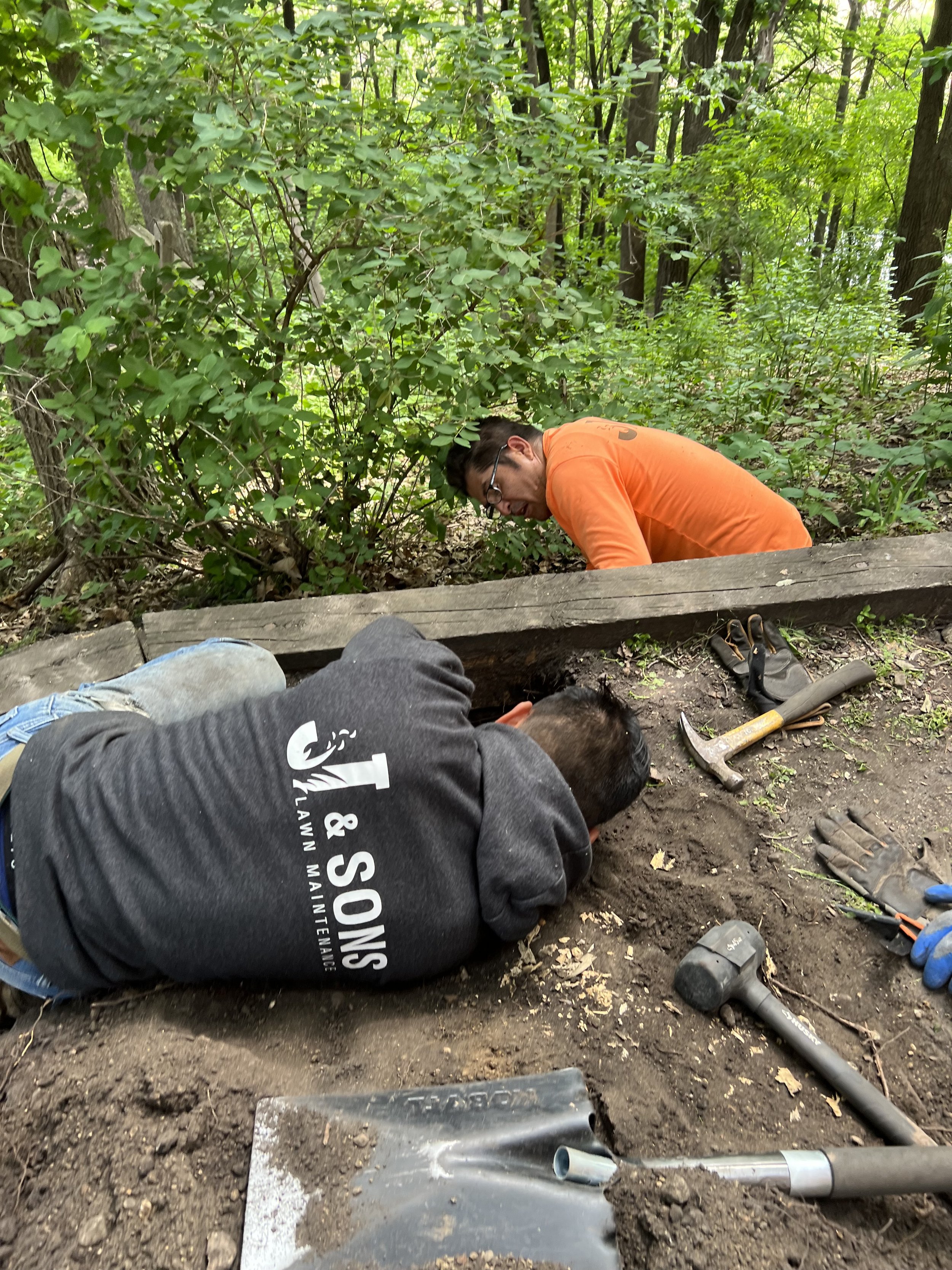 Two men working outdoors on a dirt ground near a wooden beam in a forest, using tools like a hammer and gloves, with trees and green foliage in the background.