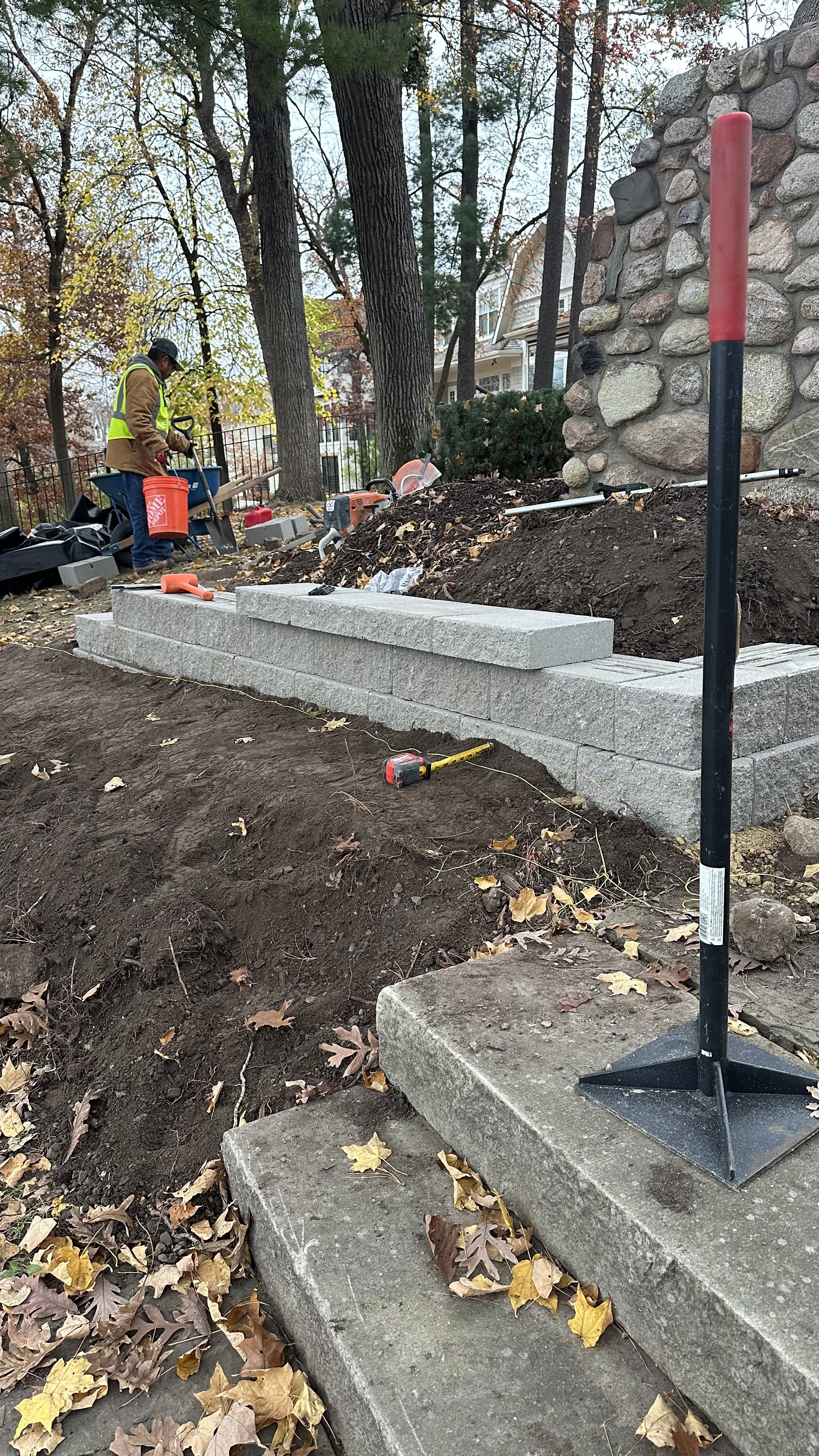 A construction worker is building a stone wall outside, with tools and materials like cinder blocks, a measuring tape, and a level nearby. The area is surrounded by trees and fallen autumn leaves.