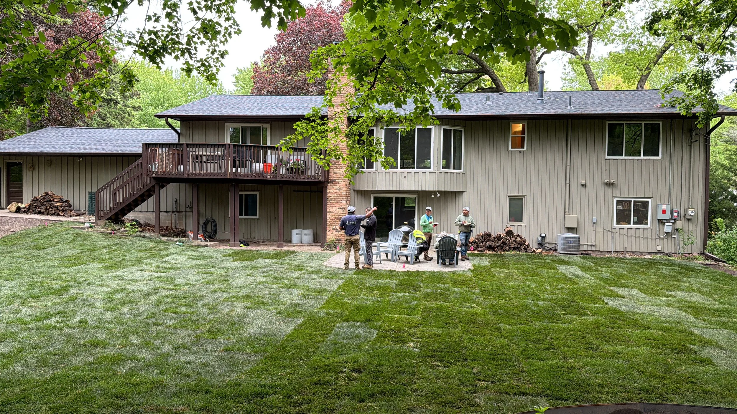 Garden or backyard under construction with people working, trees, and a house in the background.