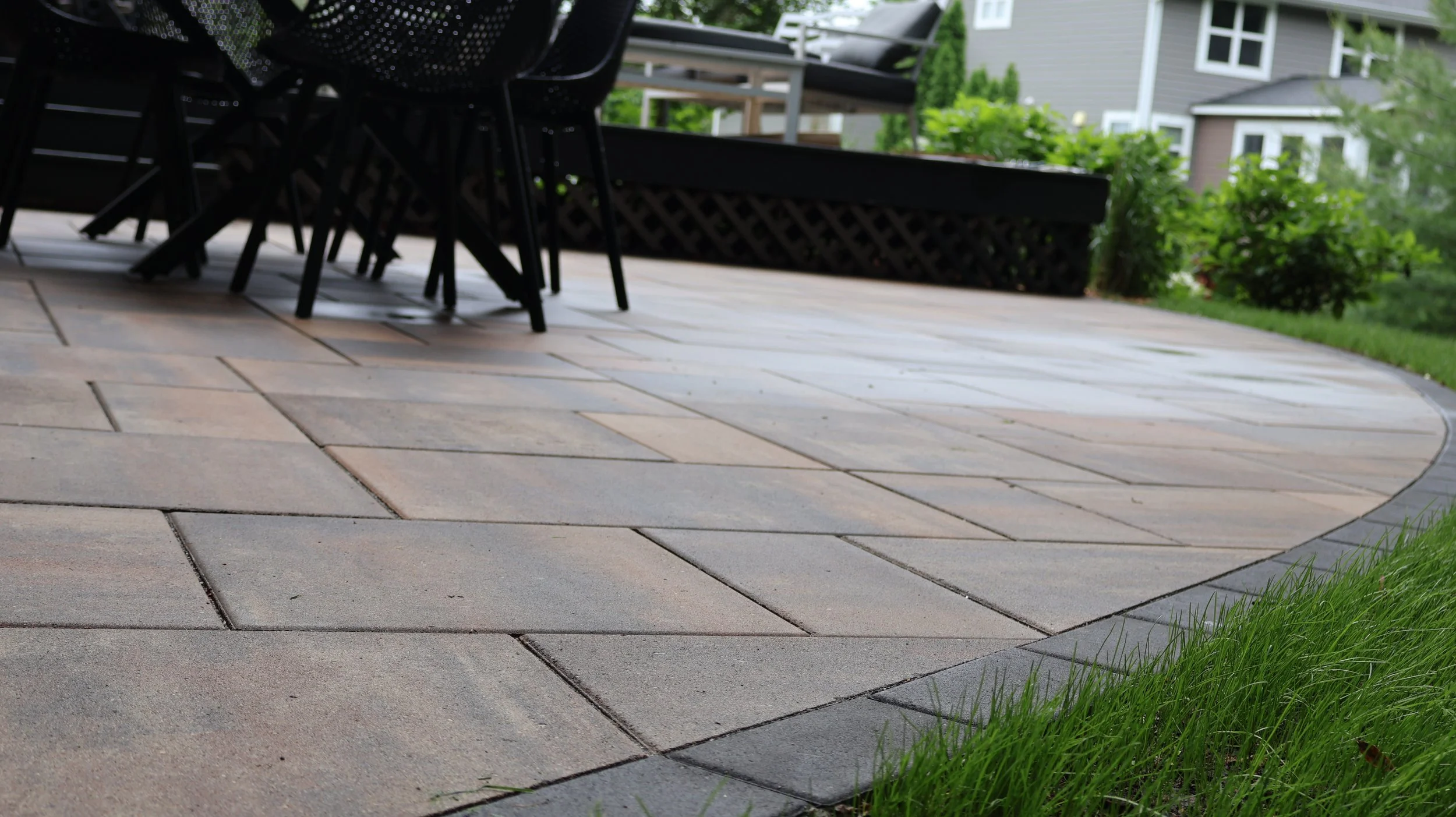 View of a patio with square pavers and outdoor furniture, including chairs and a table, surrounded by greenery and adjacent houses in the background.