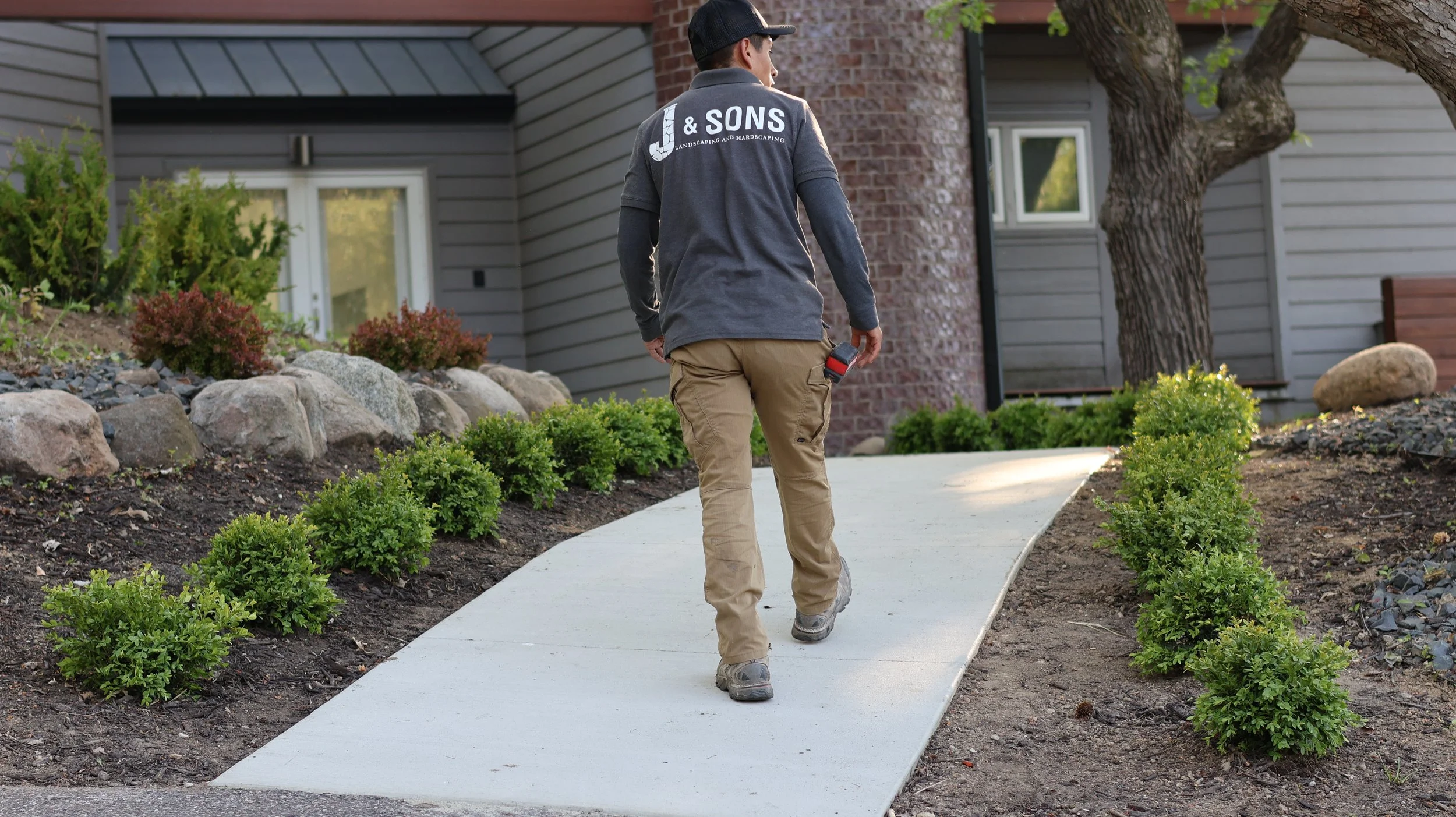 A man walking on a freshly paved sidewalk next to a landscaped garden with small bushes and rocks. He is wearing a gray jacket with ' & SONS Landscaping and Hardscaping' on the back, khaki pants, and a black cap. The house has gray siding and brick a